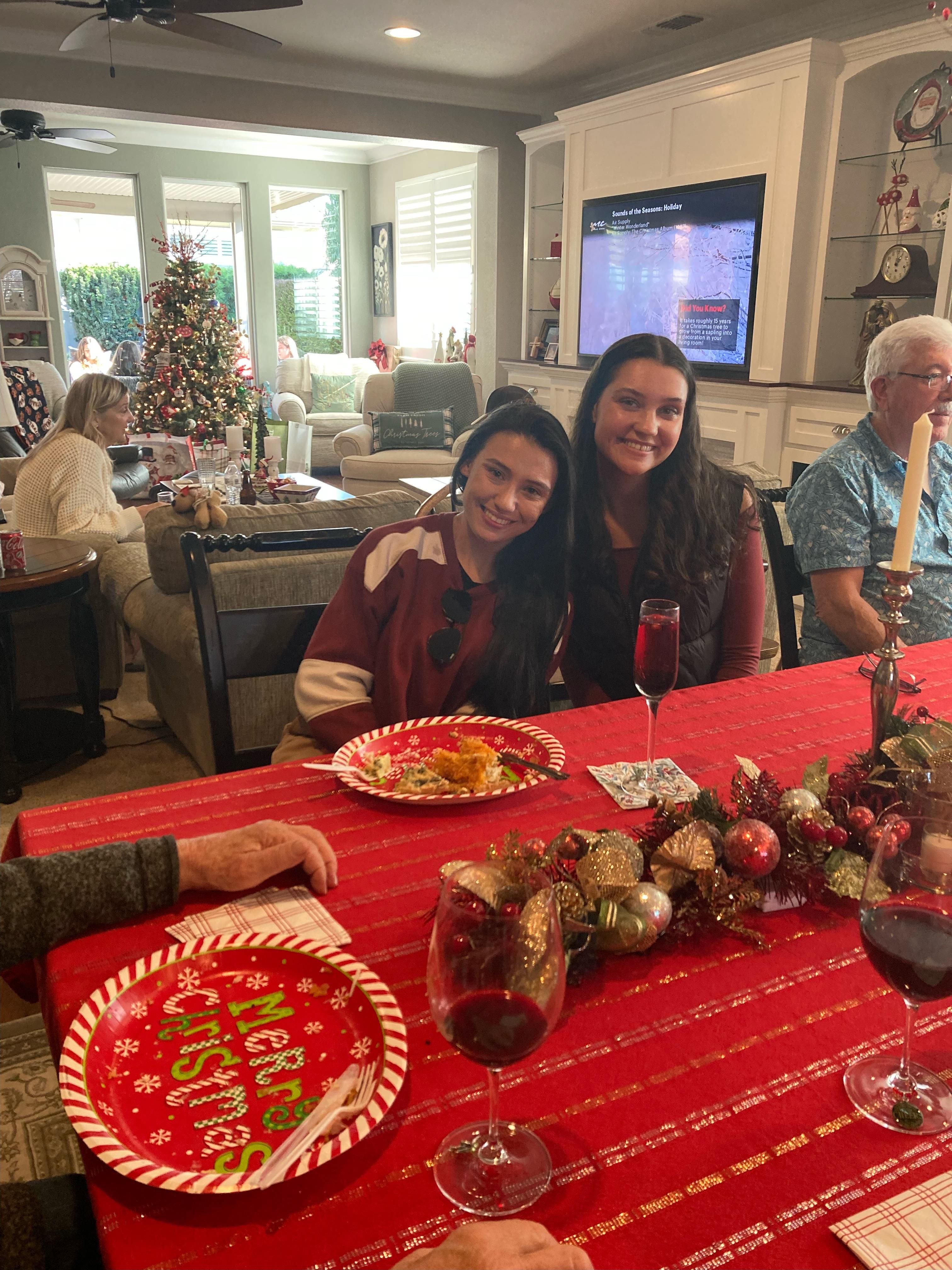 Two friends smile at the camera while sharing a festive meal in a decorated living room.