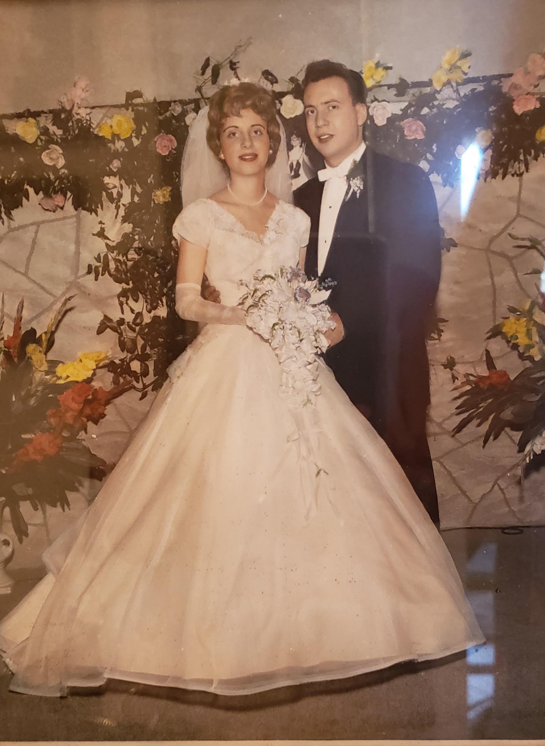 Couple dressed in formal wedding attire poses with floral arrangements in the background.