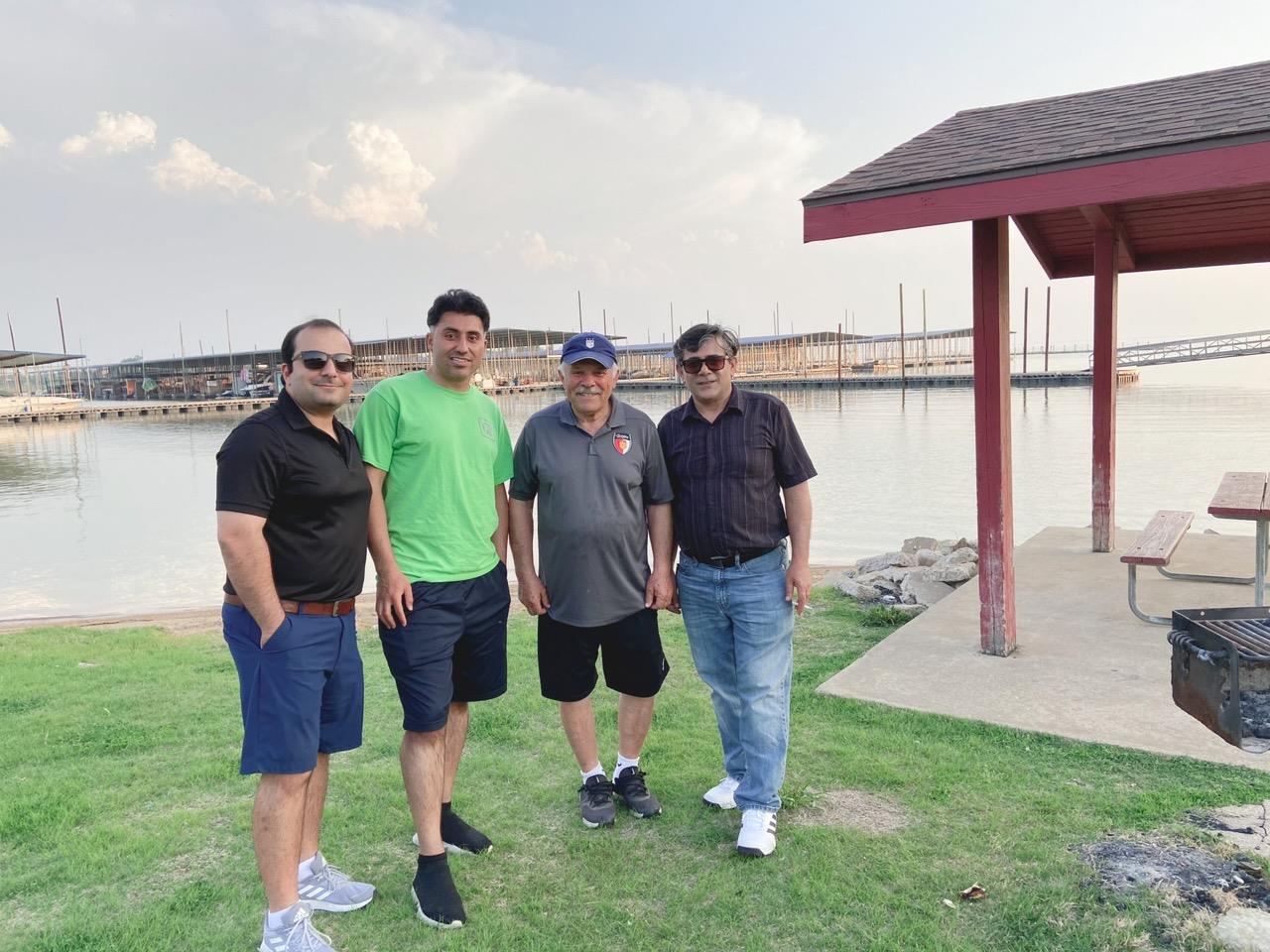 Four friends stand together on the grass by the lake, enjoying their time in the sunshine.