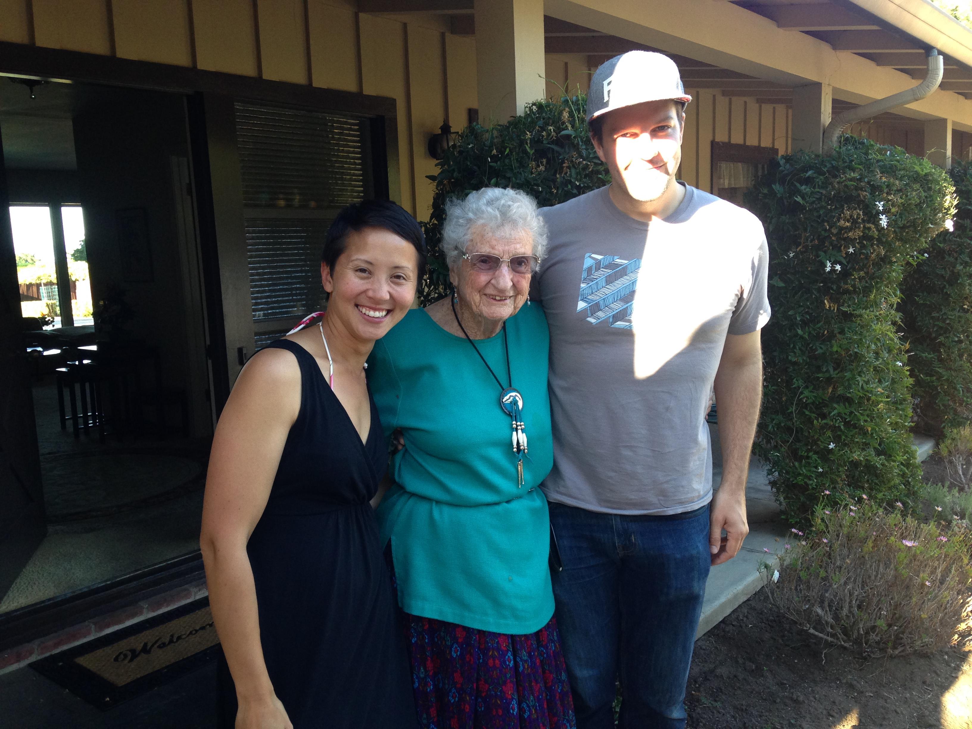 Three people smile together outside a charming house on a bright, sunny day.