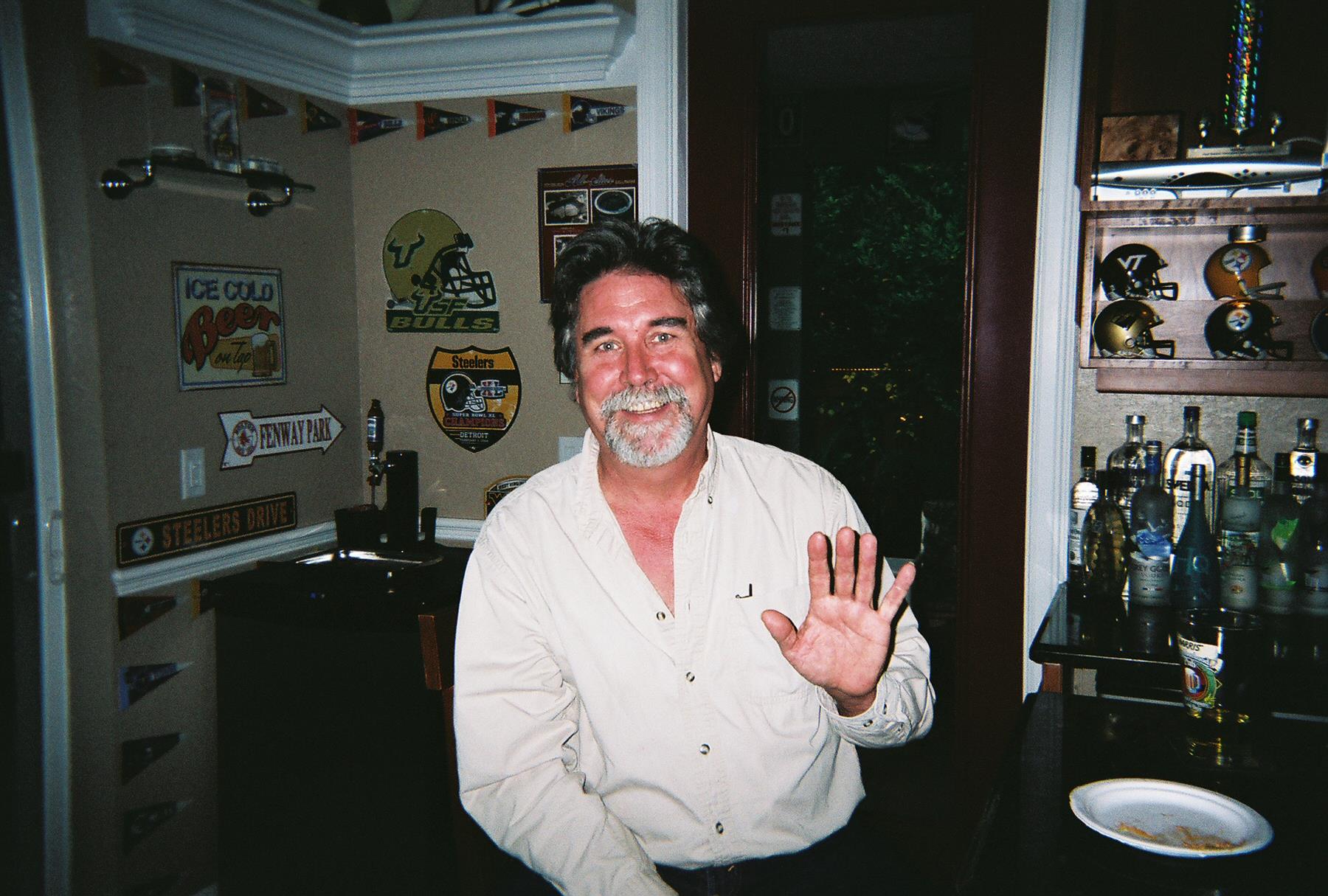 A man with a beard greets with a wave in an inviting room filled with decor and collectibles.