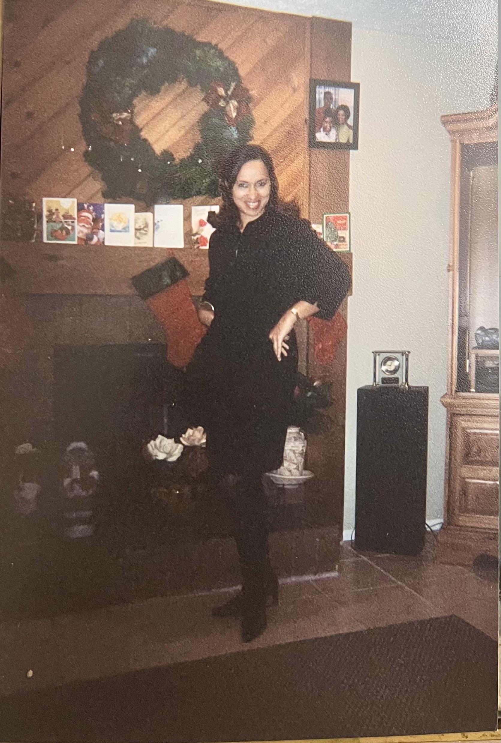 A woman joyfully poses by a festive fireplace, displaying her elegant outfit and holiday cheer.