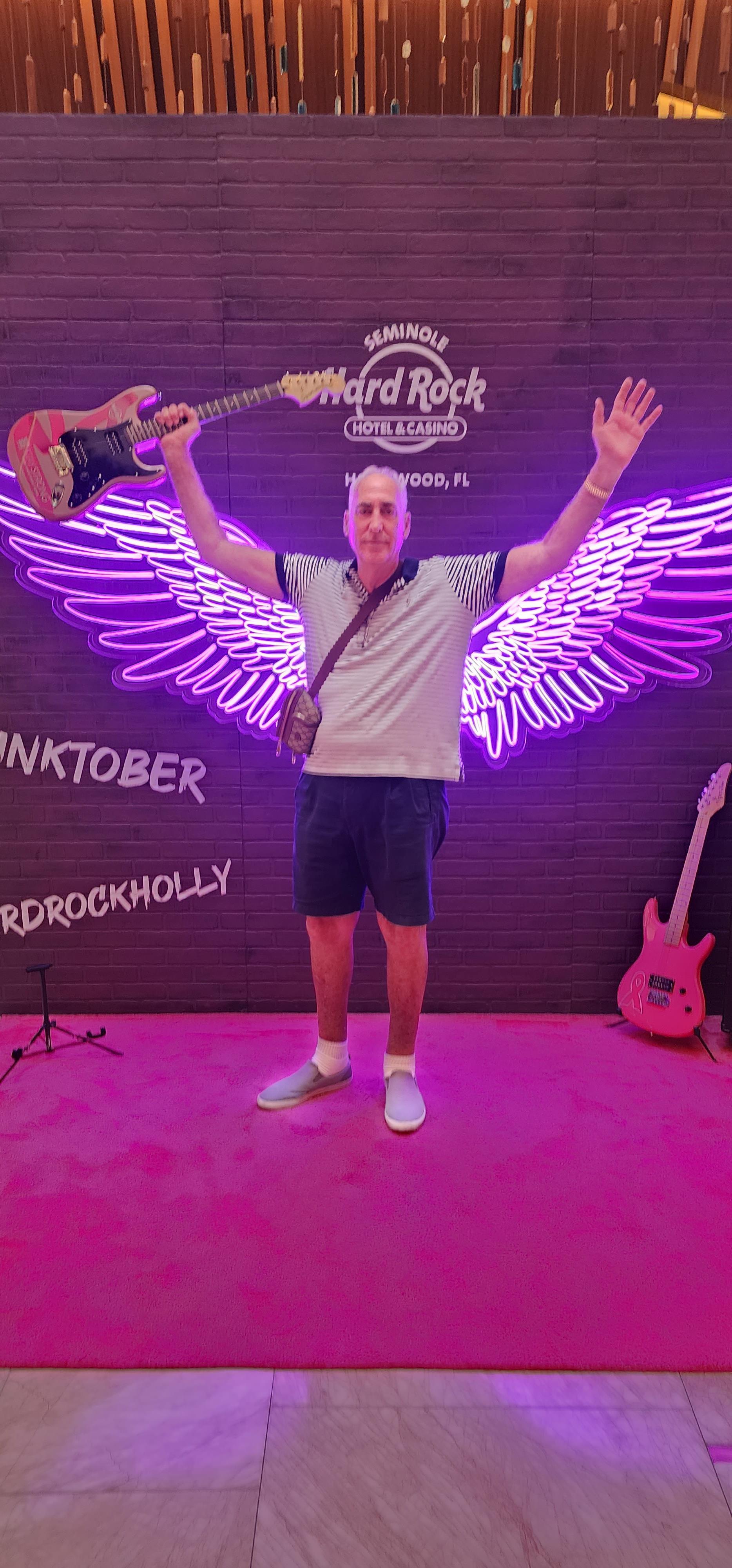 A man celebrates at a music festival, posing joyfully with a guitar by neon wing art.