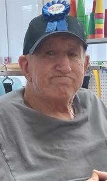 An elderly man sits indoors, wearing a cap and gray shirt, displaying a thoughtful expression.
