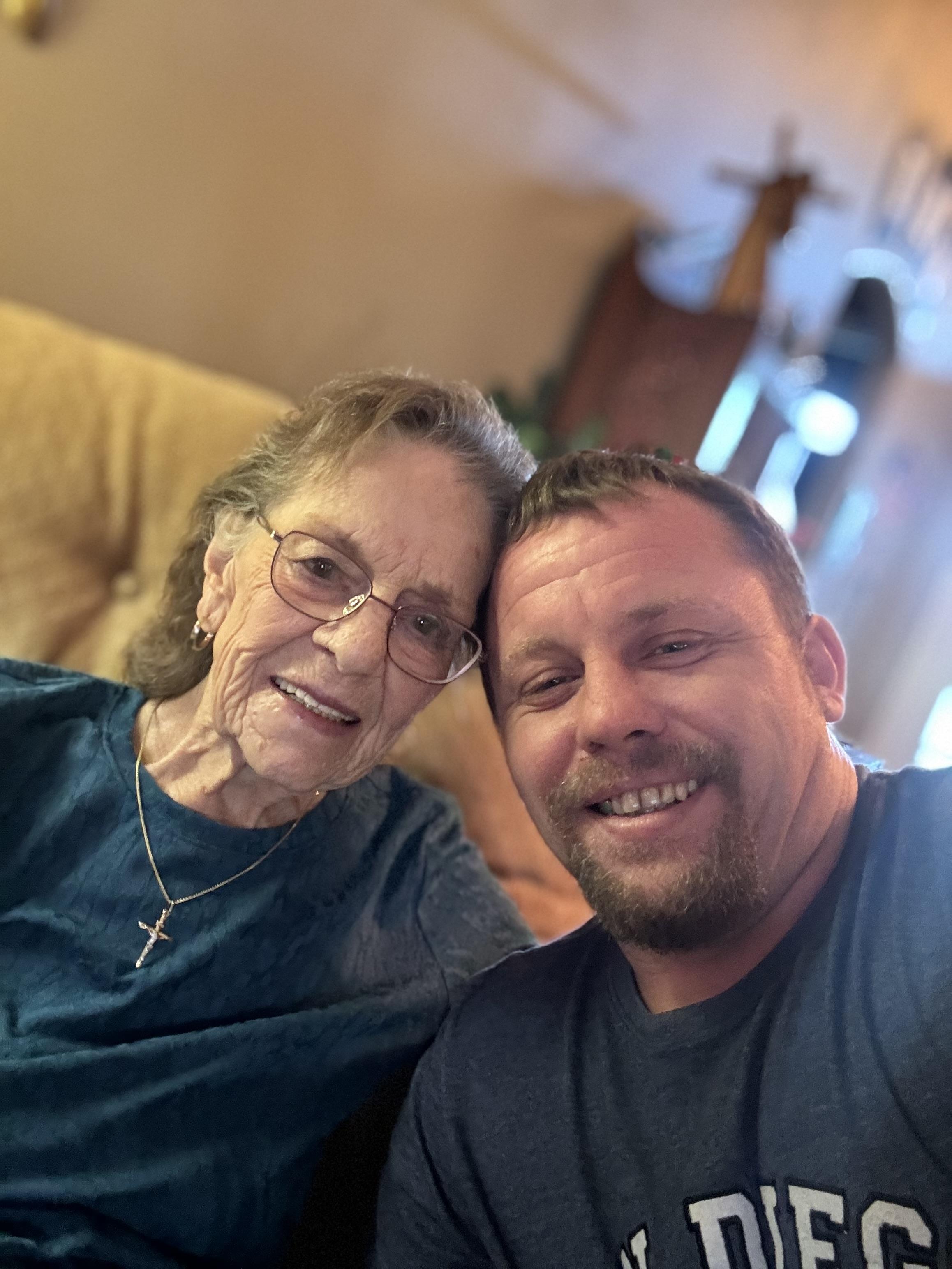 A grandmother and her grandson share a warm smile while sitting together on a couch in their home.