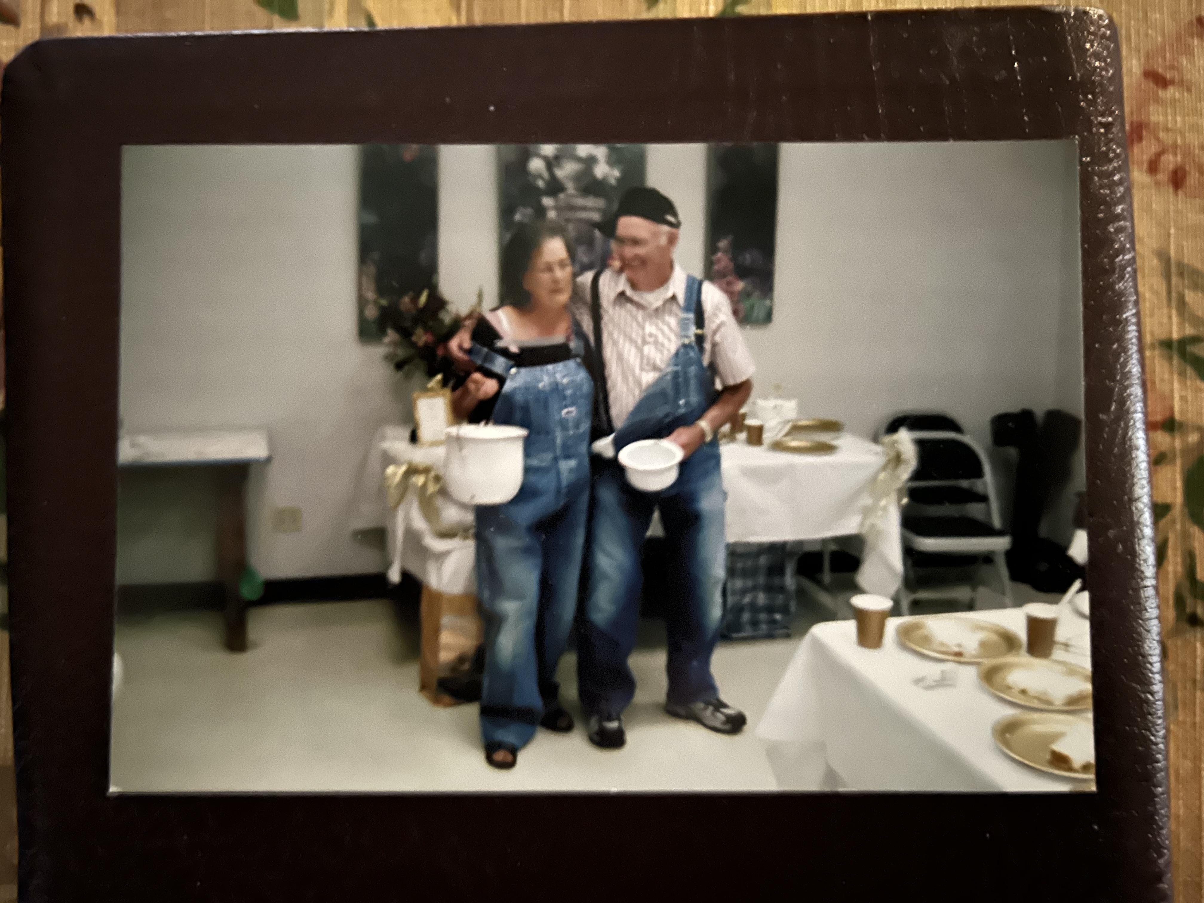 A cheerful couple stands together, holding food containers at a lively gathering with tables.
