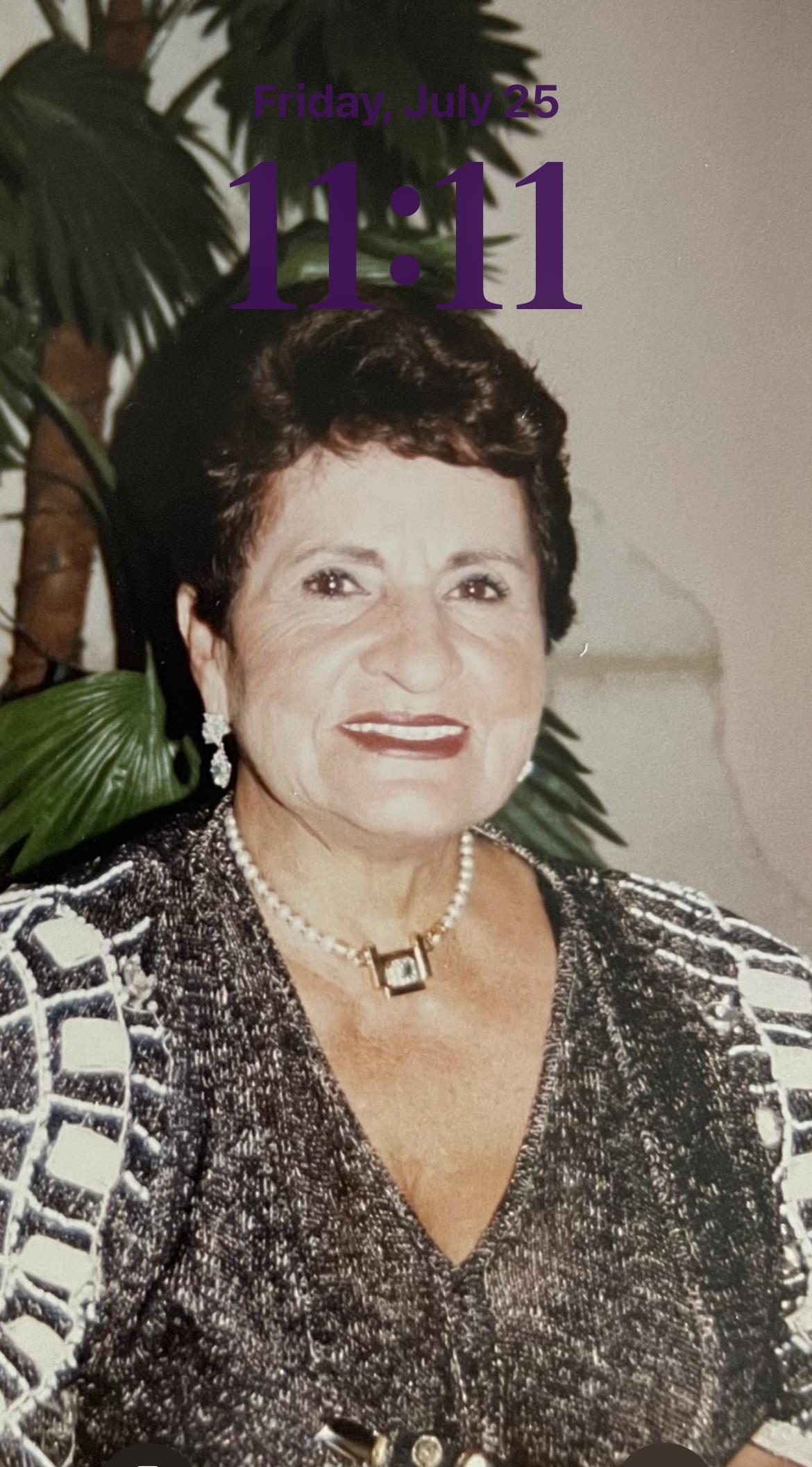 Woman dressed in elegant attire, smiling warmly at a formal event surrounded by plants.