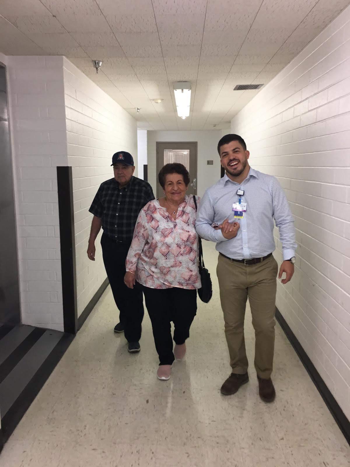 Two people walk in a bright hallway, enjoying time with a young man in a formal shirt.