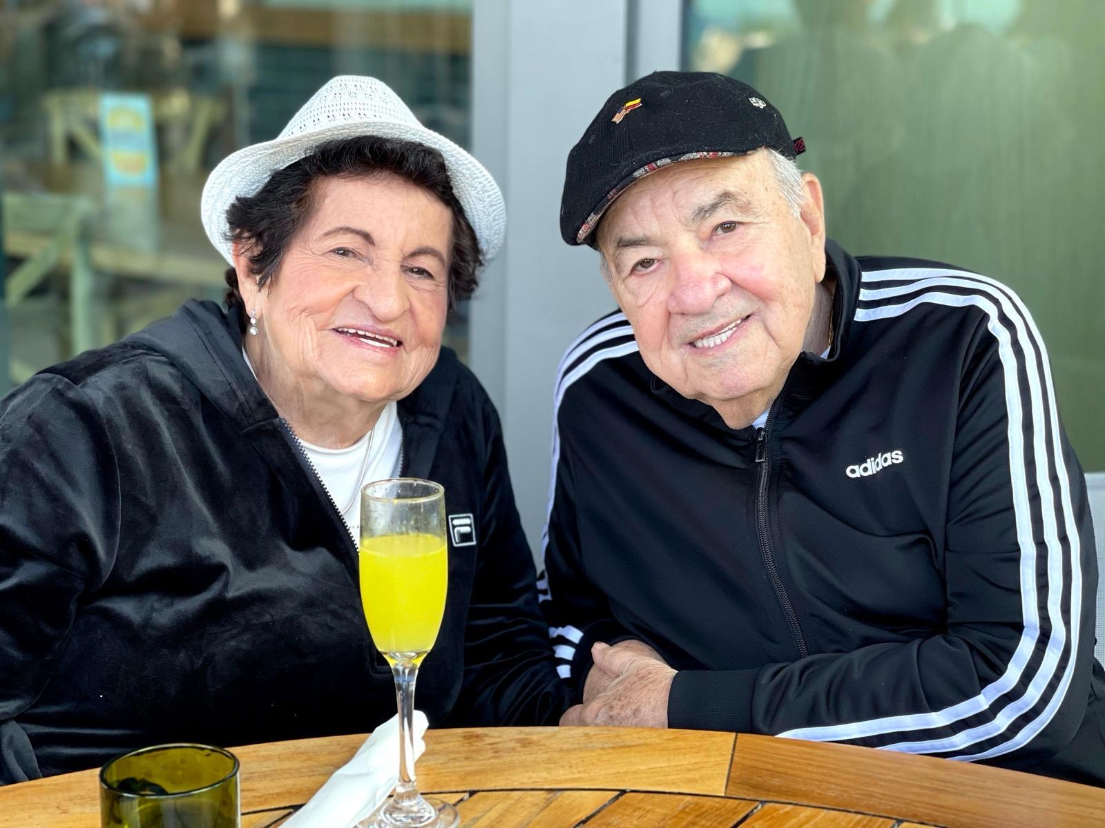 Two older adults smile at the camera while sharing a drink in a casual cafe setting.
