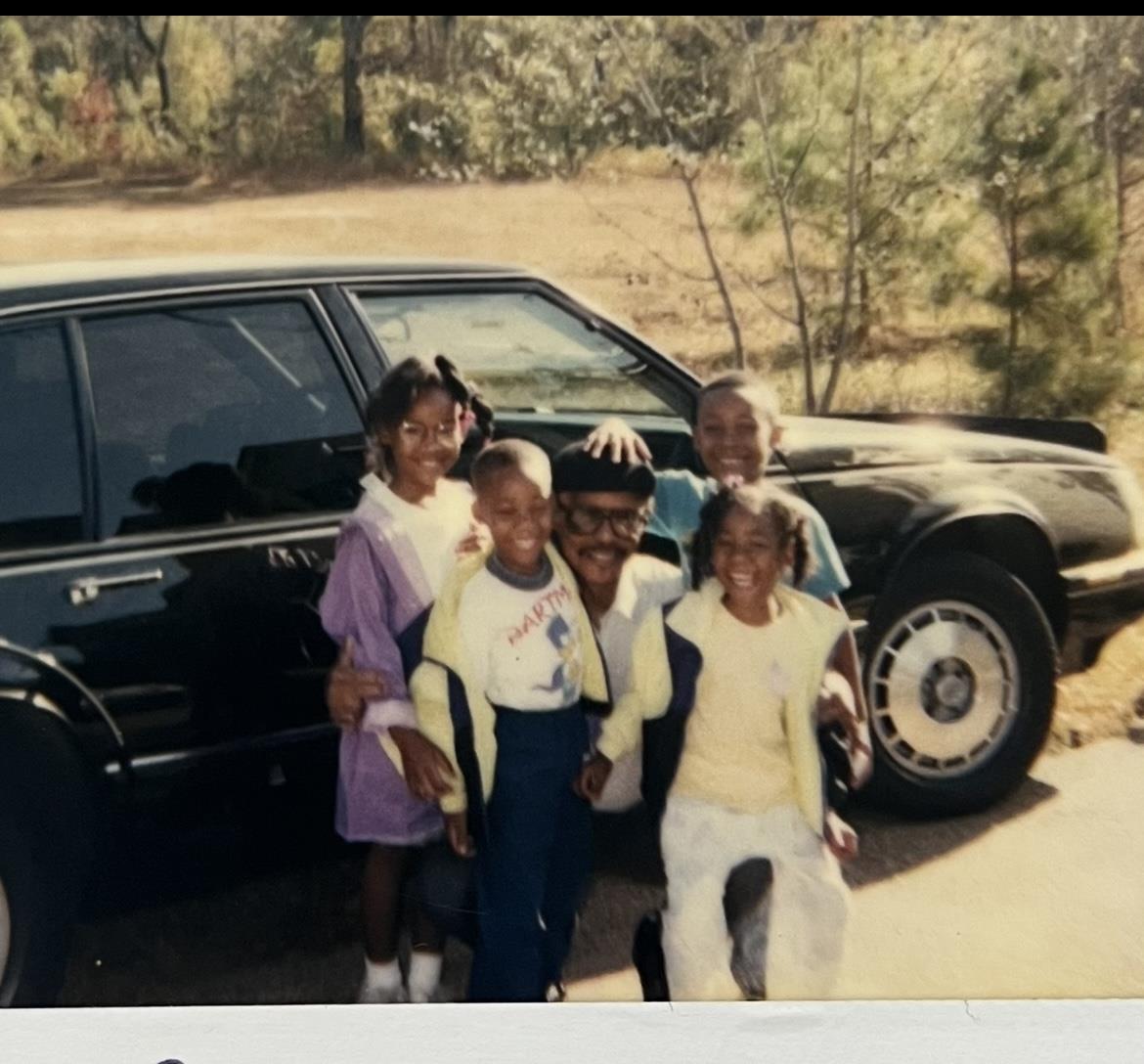 Five children smile and pose together by a black car in a grassy area on a sunny day.