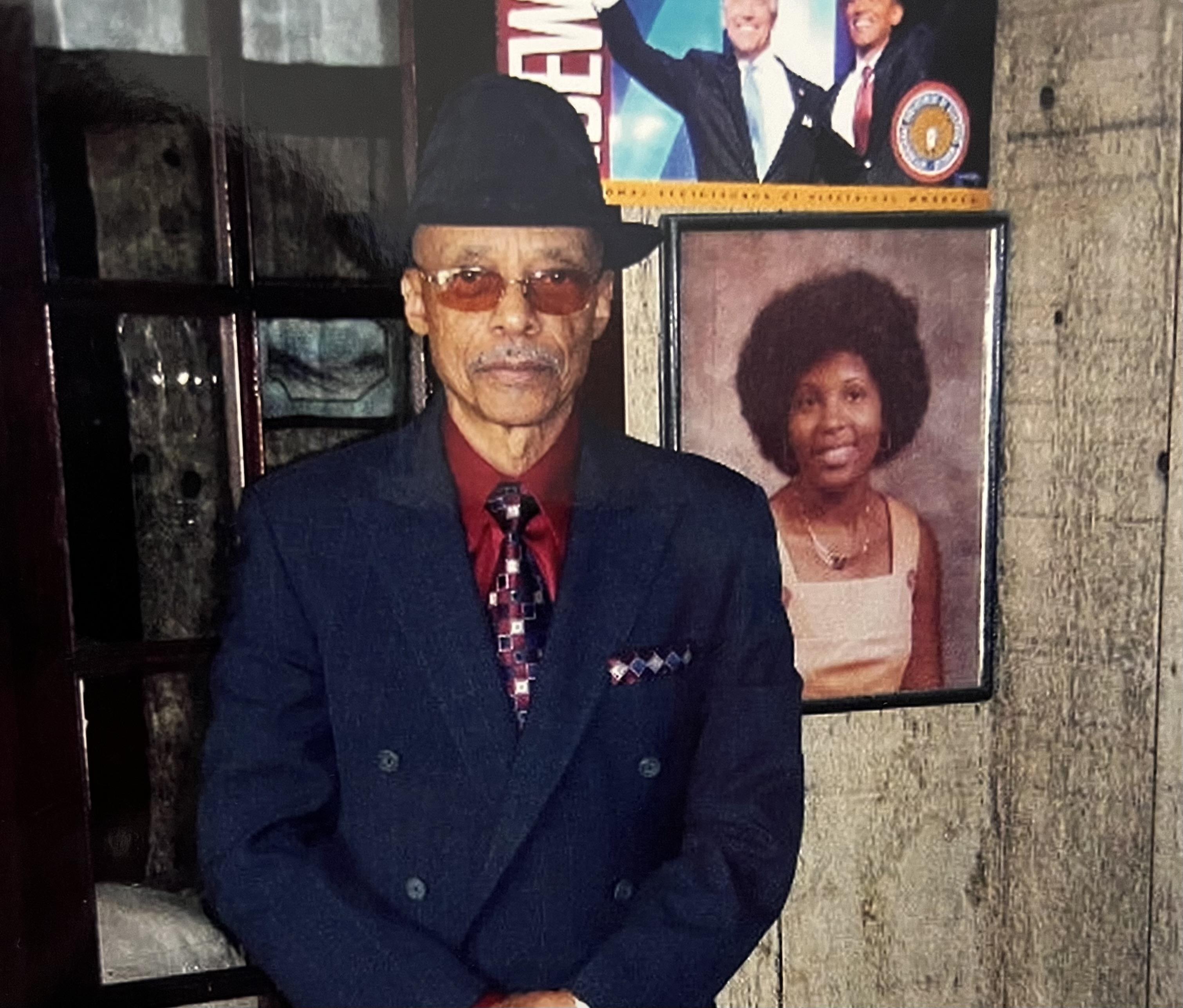 Elderly gentleman in formal attire stands confidently near historical portraits on the wall.