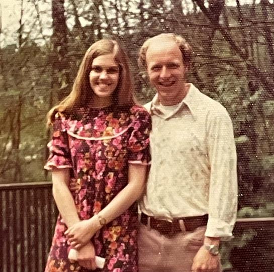 A couple stands close, smiling joyfully in a lush forest during spring, dressed in vintage attire.