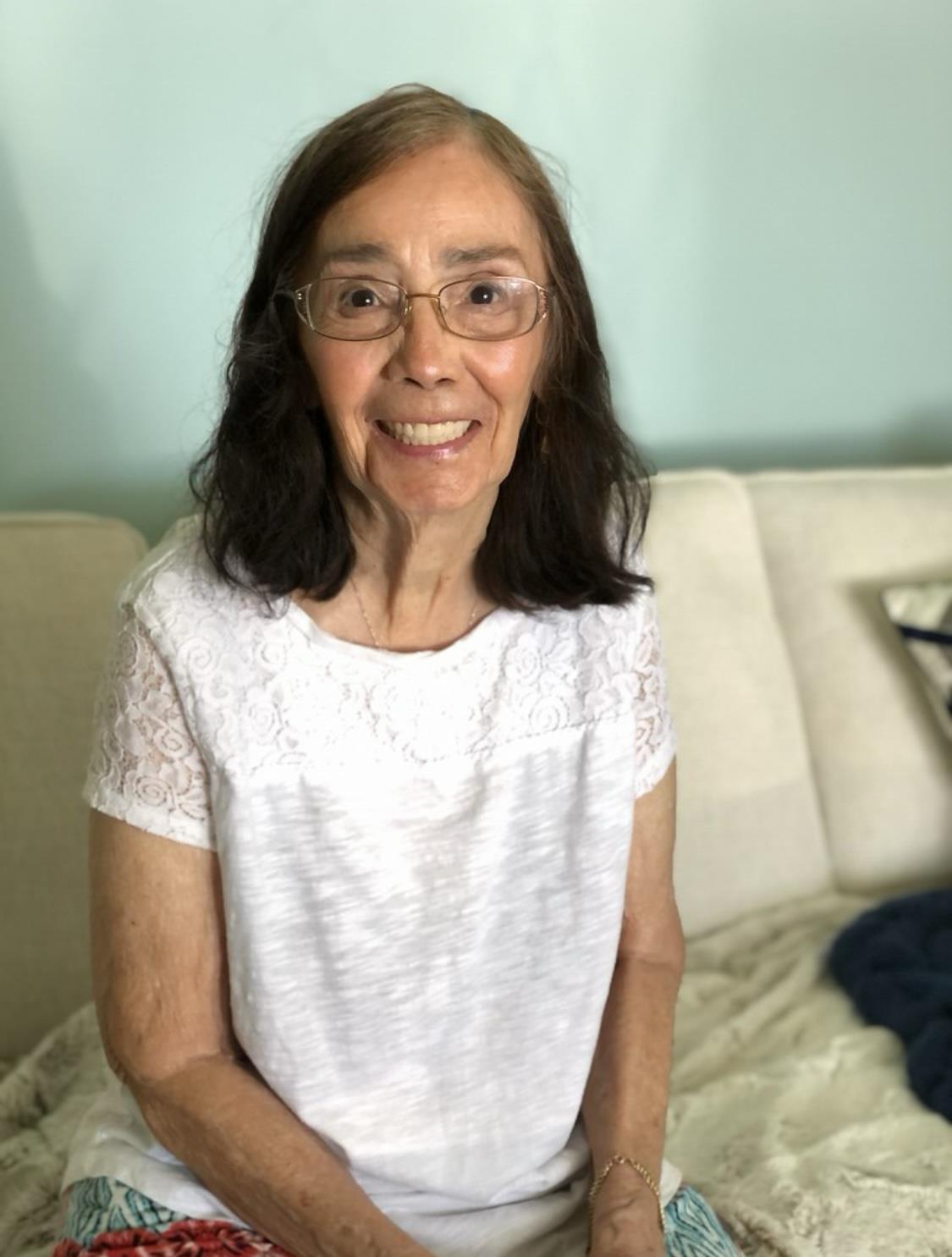 A cheerful elderly woman with glasses sits on a couch, smiling warmly in a cozy room.
