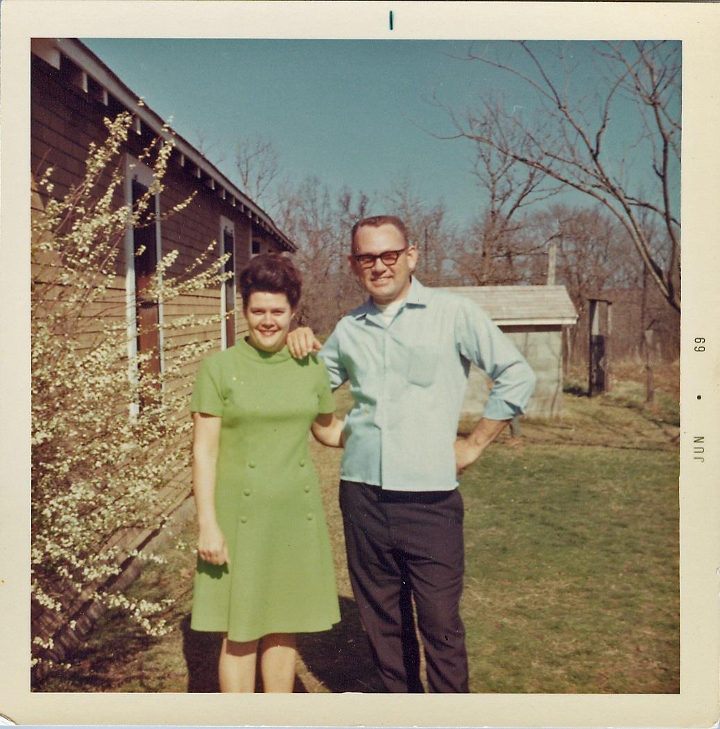 A couple stands in their backyard, smiling and enjoying a sunny day surrounded by blooming trees.