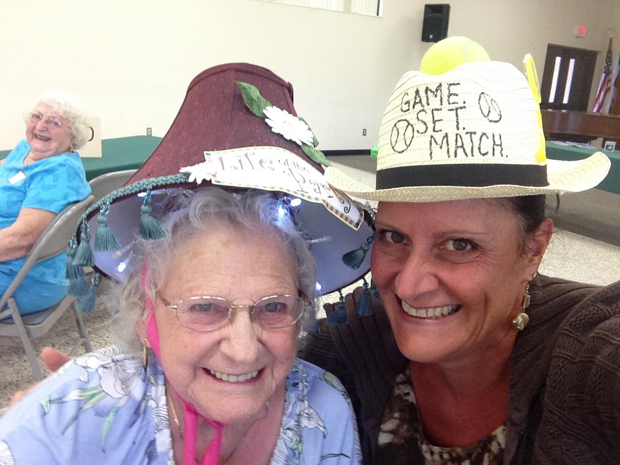 Two women joyfully wear creative hats at a community event, showcasing togetherness.