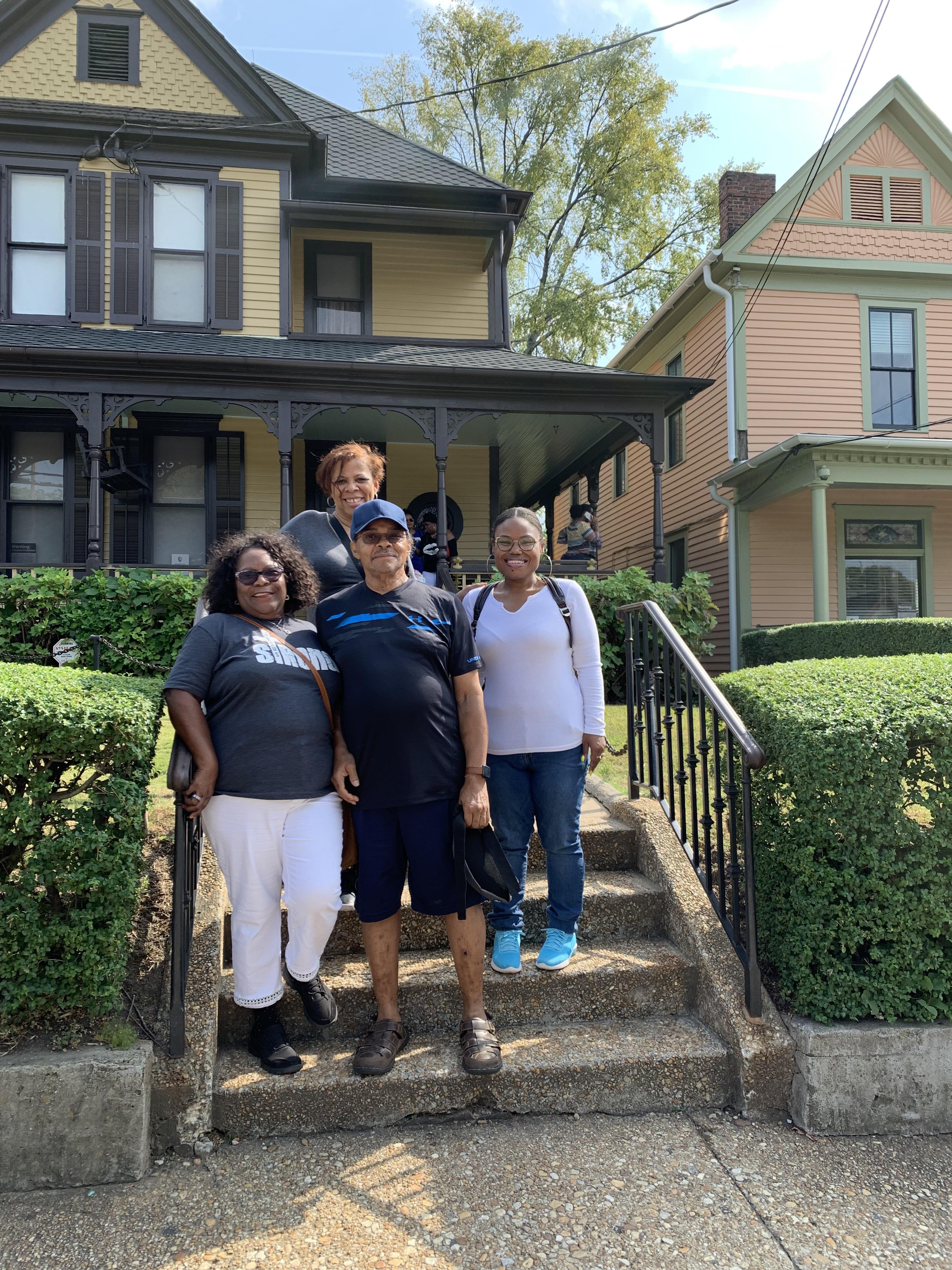 Four family members stand on the steps of a well-maintained historic house, smiling happily.