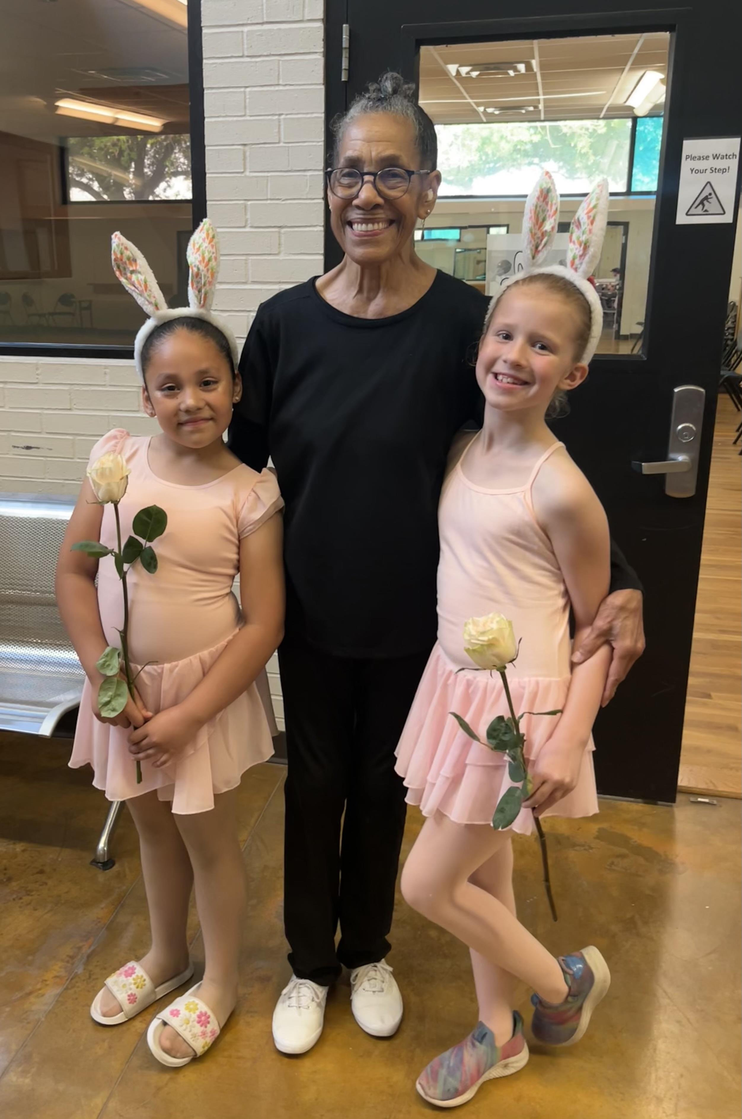 Two young ballet dancers with flower props stand beside their instructor in a dance studio.