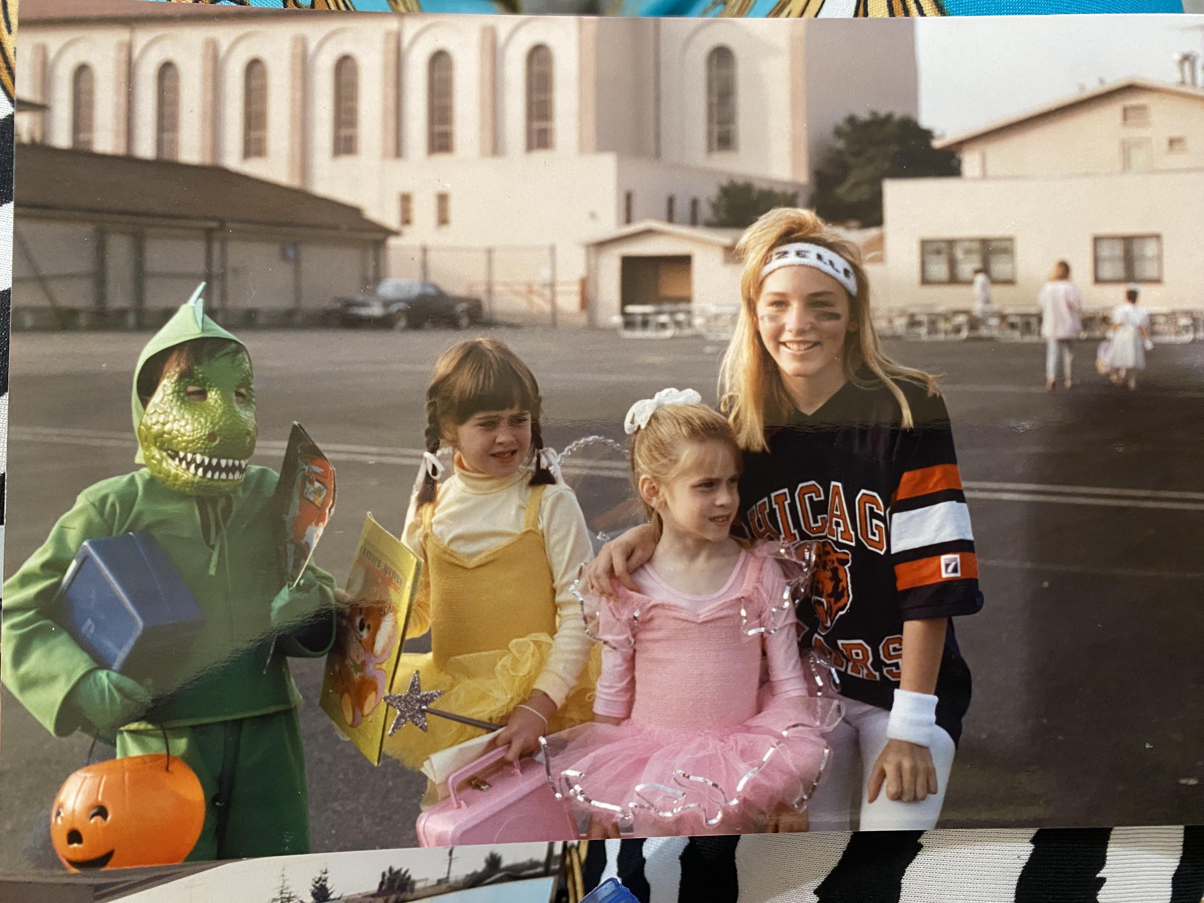 Four kids dressed in various costumes stand together happily in front of a school building.