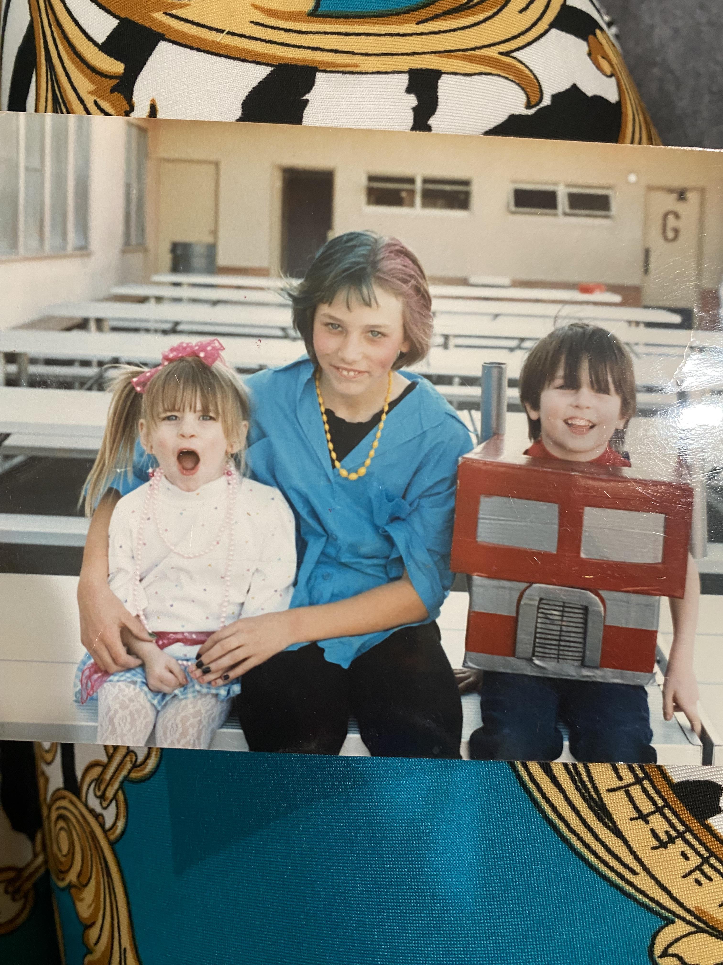Three children smile while sitting together outdoors, with one in a costume.