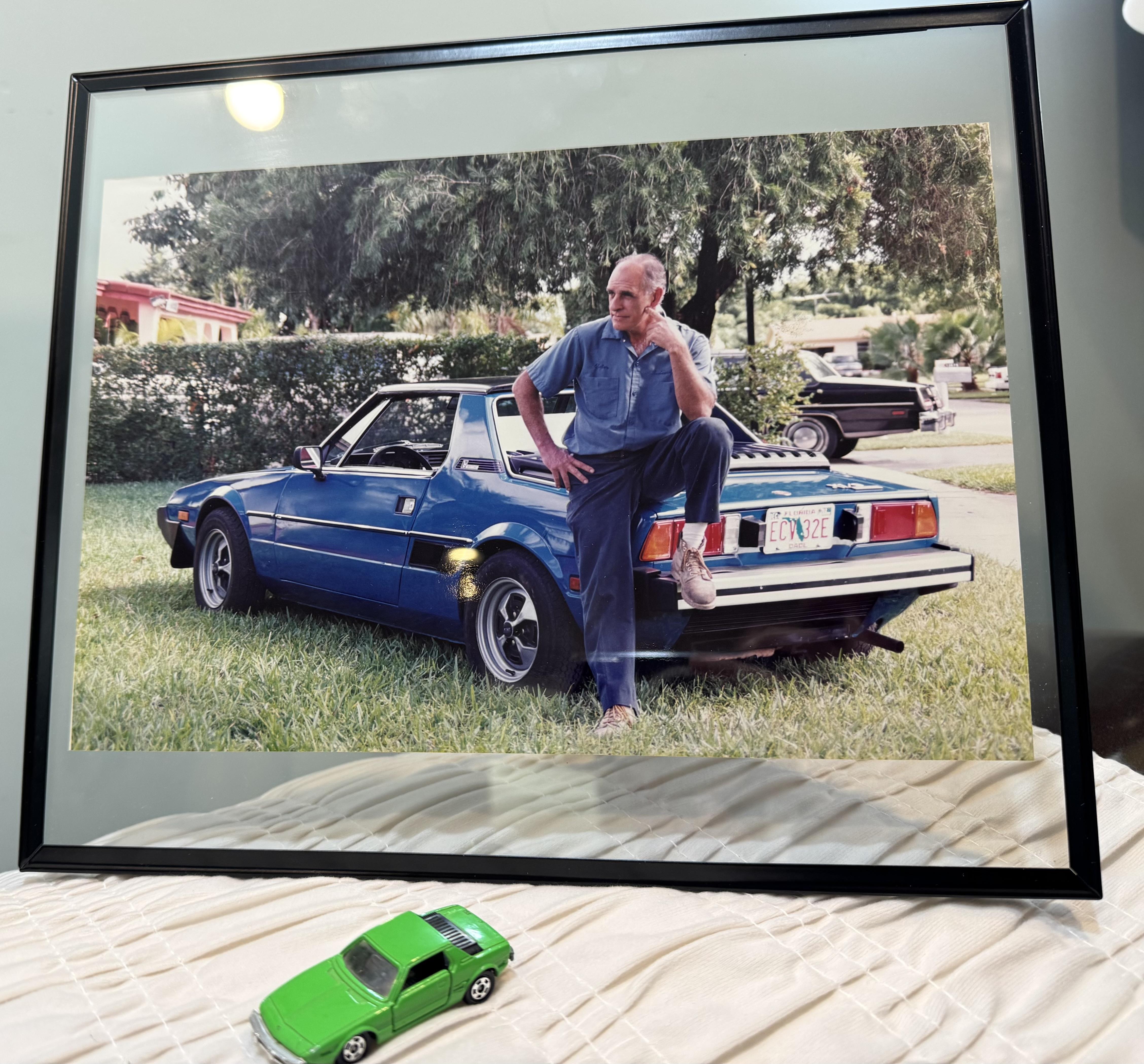 A man casually poses on a bright blue vintage car parked on green grass in sunlight.