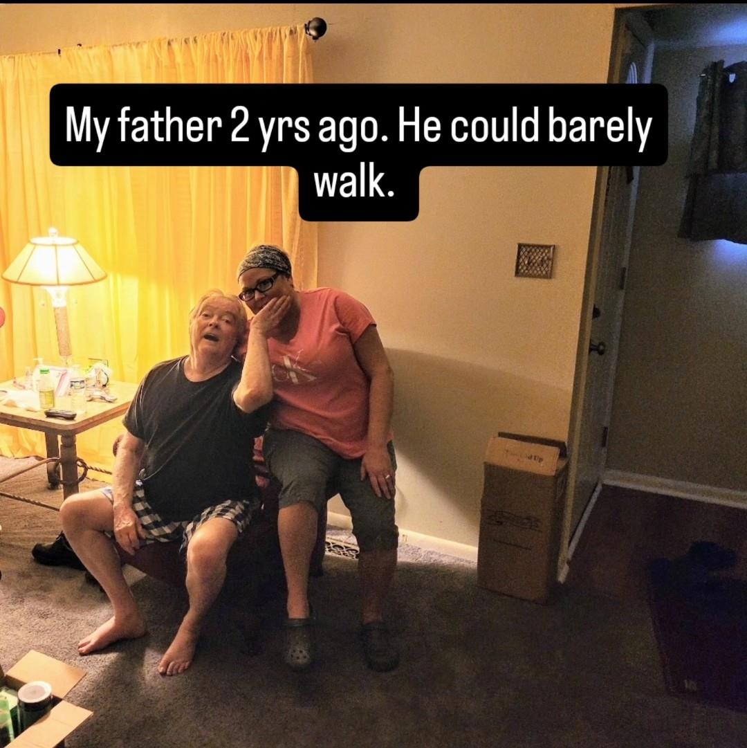 A son comforts his father, who is struggling with mobility, in their living room setting.