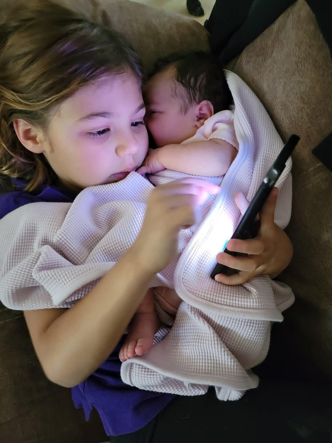 Older sibling interacts with smartphone while cradling infant sibling in cozy blanket.