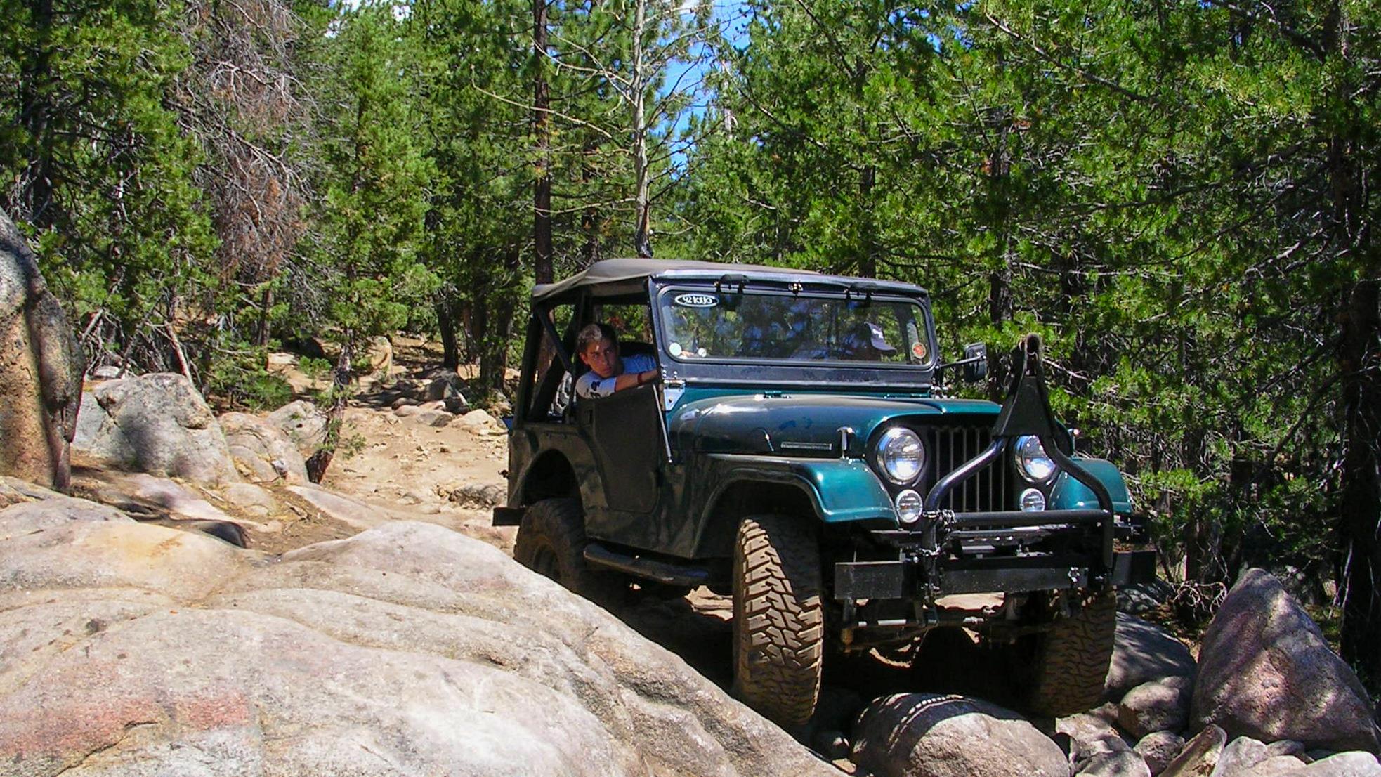 A rugged vehicle climbs over large rocks, surrounded by tall trees under a bright sky.
