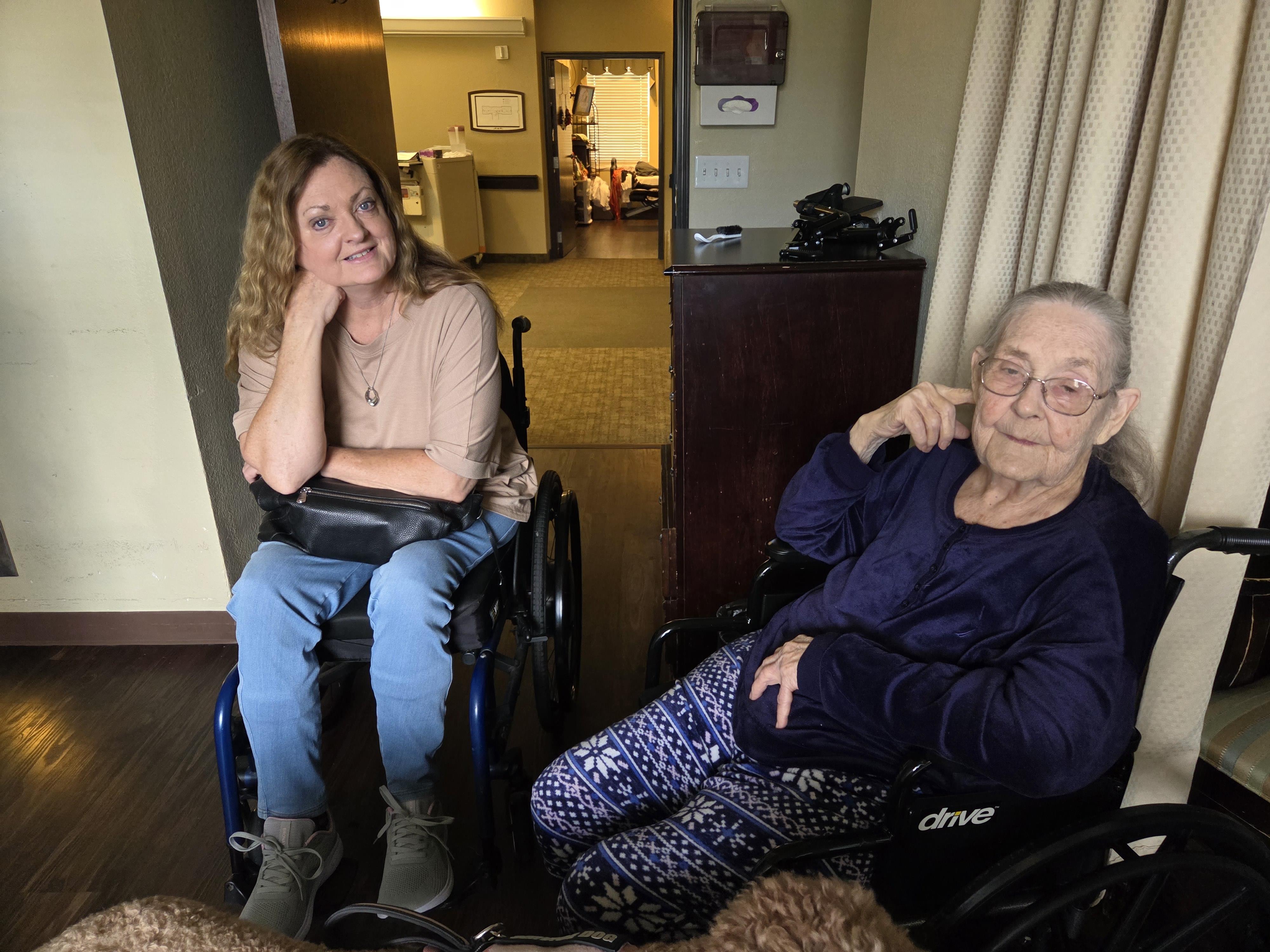 Two women sit in wheelchairs, chatting amiably in a care facility, enjoying their time together.