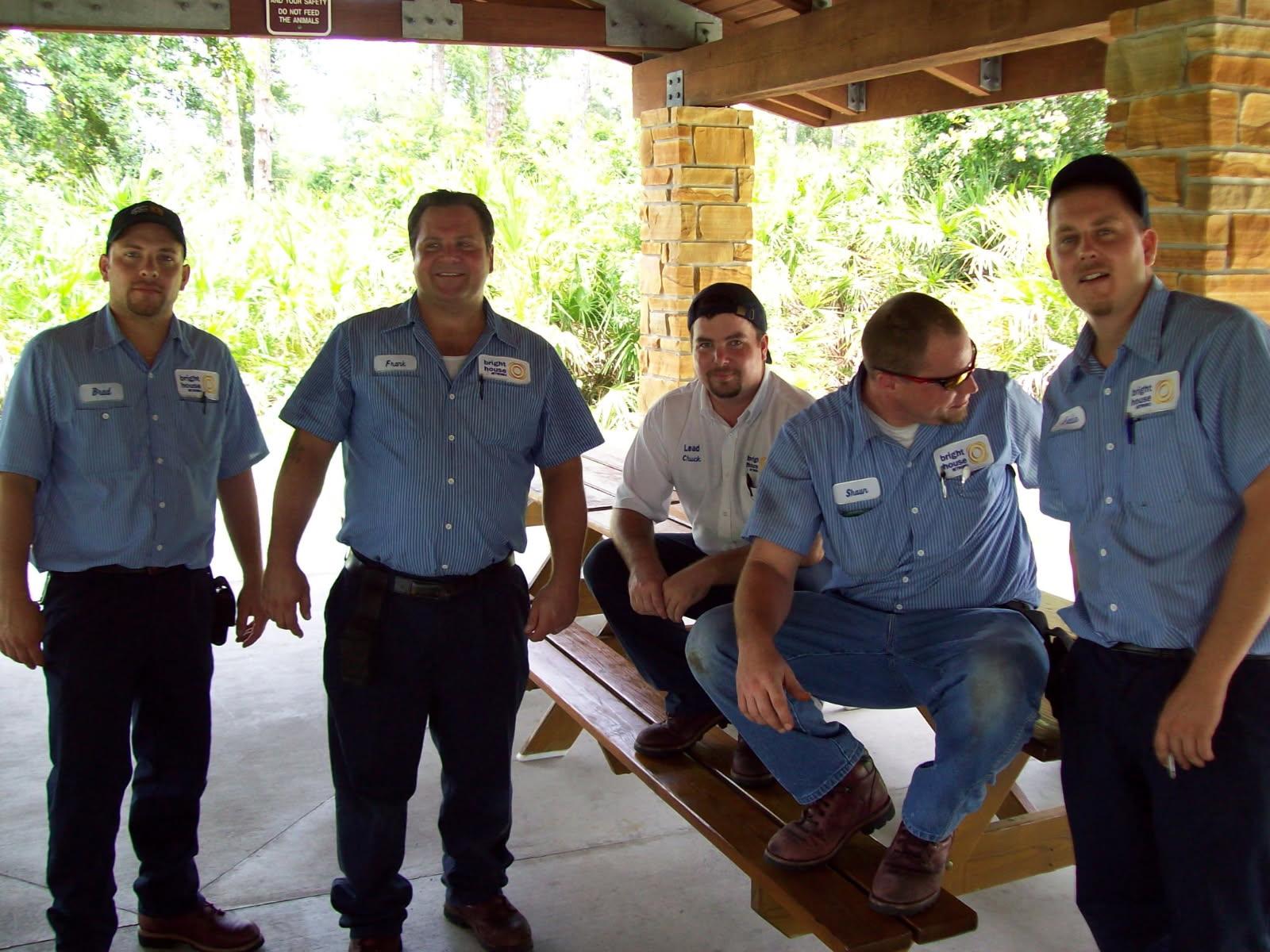Five workers in blue uniforms relax at a pavilion, engaged in conversation during a break.
