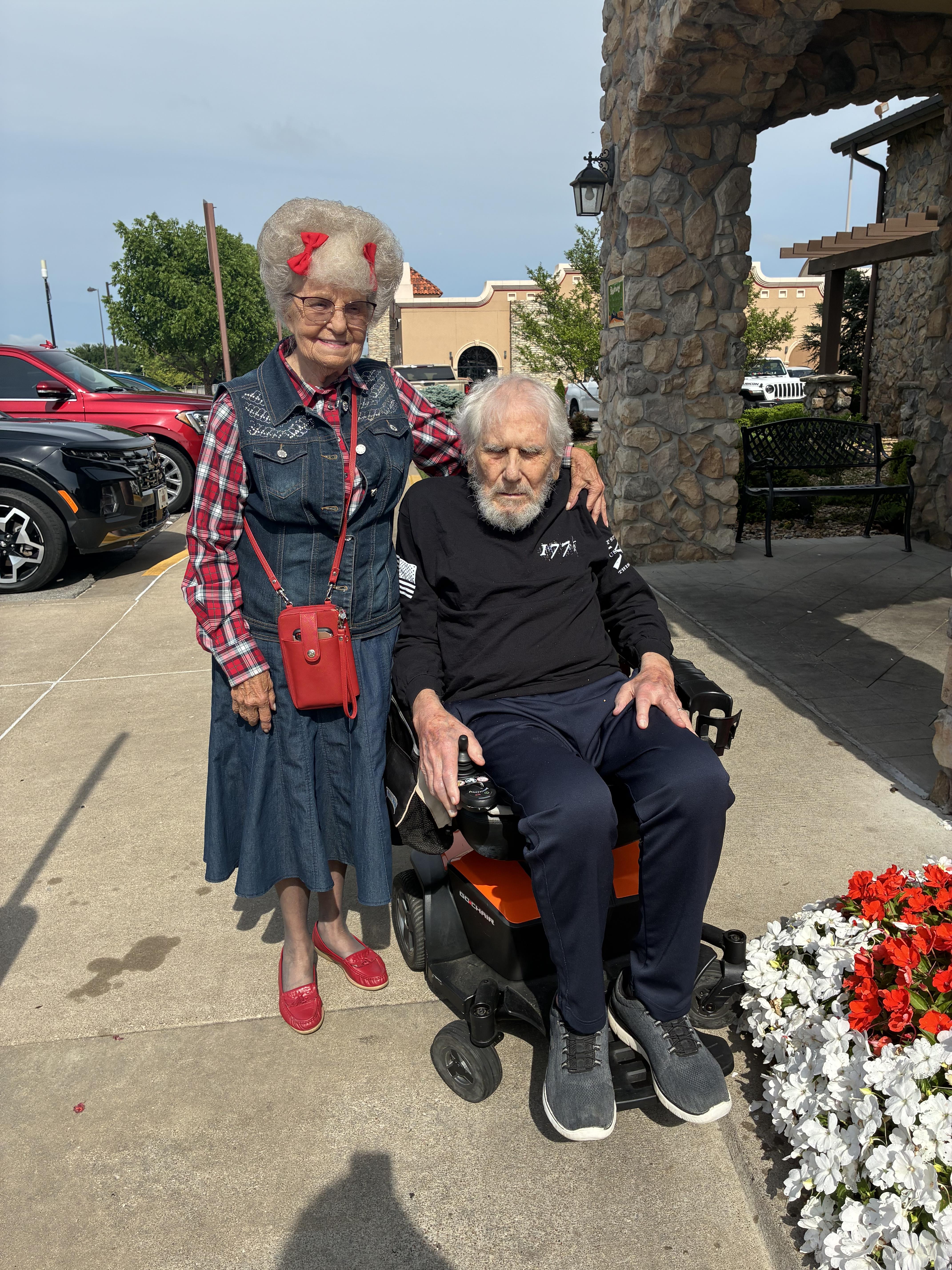 An elderly couple poses together outdoors, enjoying time in the sun with bright flowers nearby.