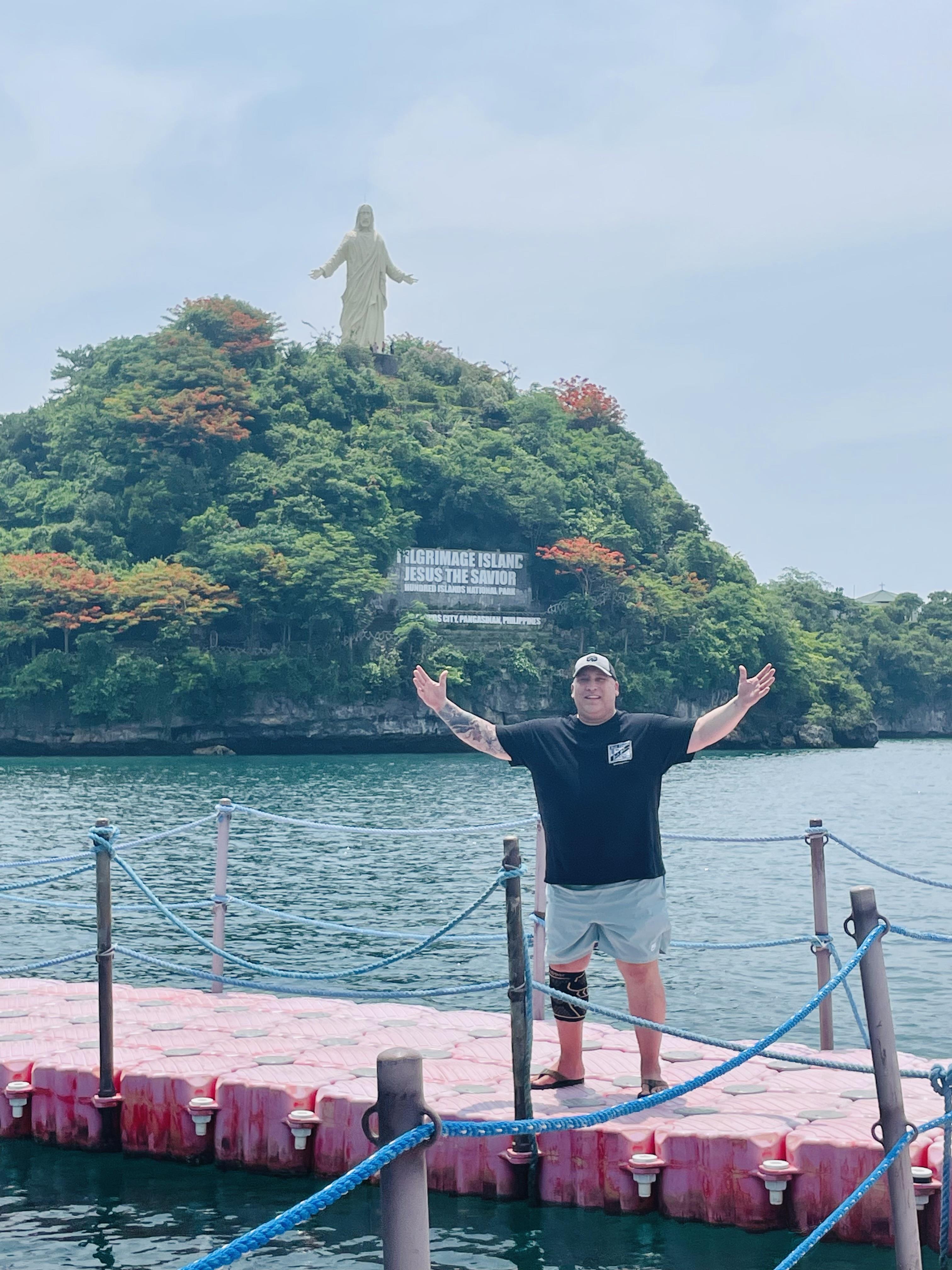 A person stands with arms open in front of a statue on a lush island, enjoying a sunny day.