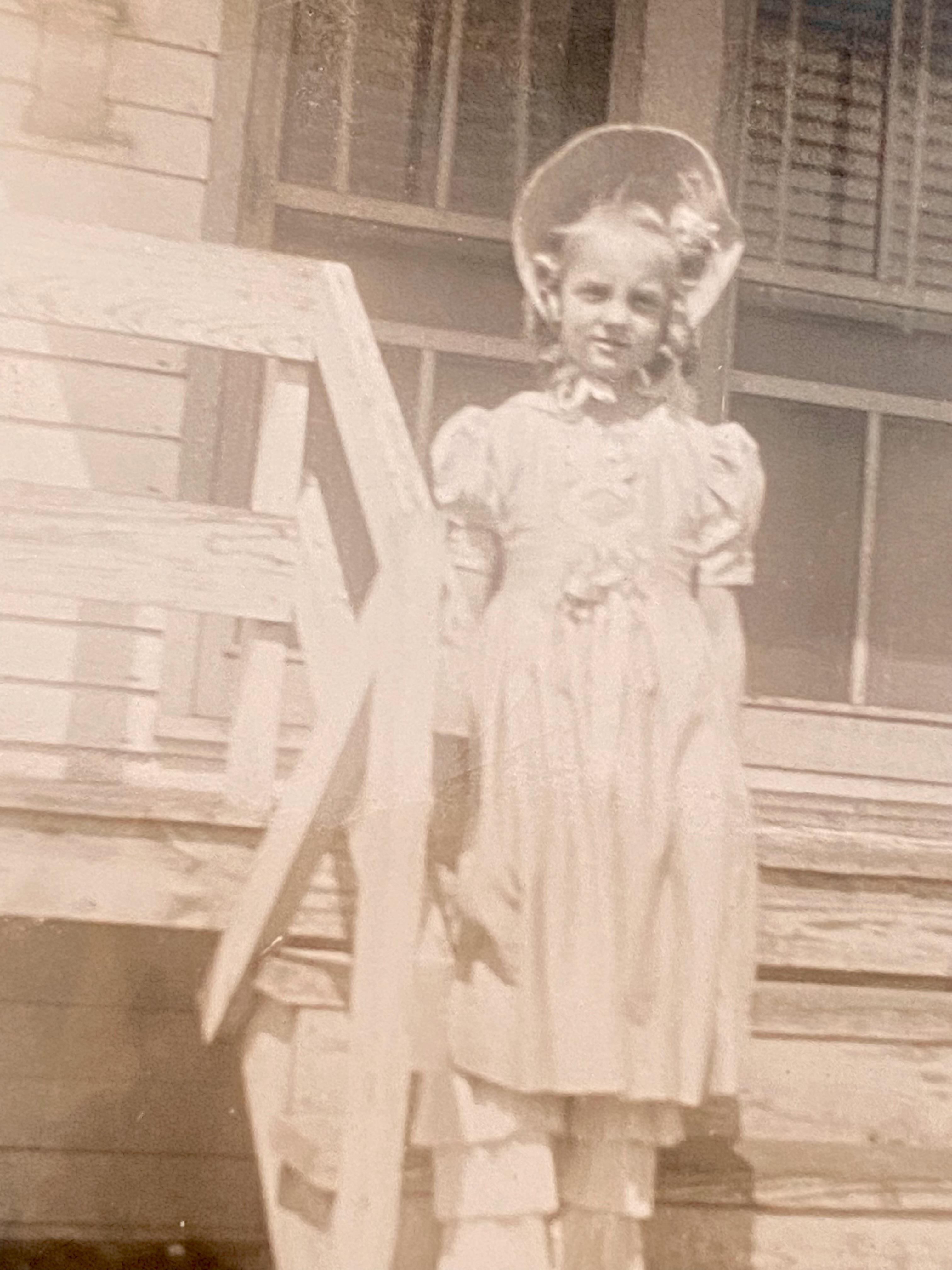 A young girl wearing a light-colored dress poses on the steps of a farmhouse with a smile.