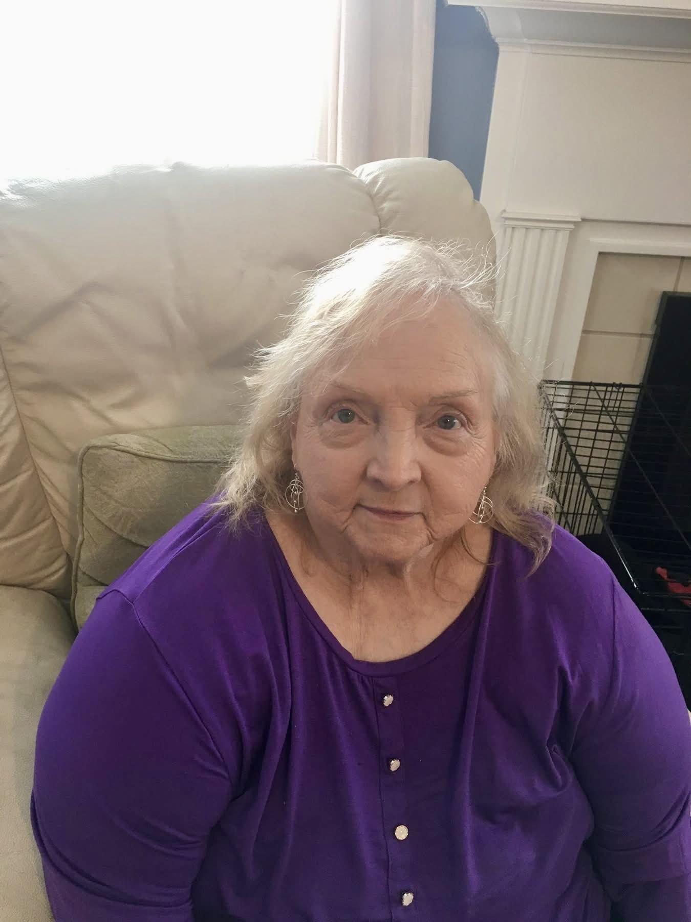 Older woman with silver hair is seated on a light-colored couch in a comfortable living room.