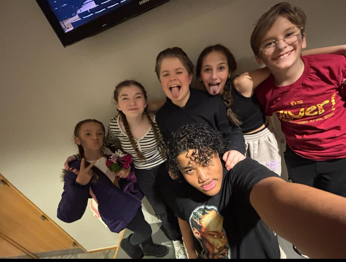 A group of six kids poses cheerfully together while taking a selfie indoors.