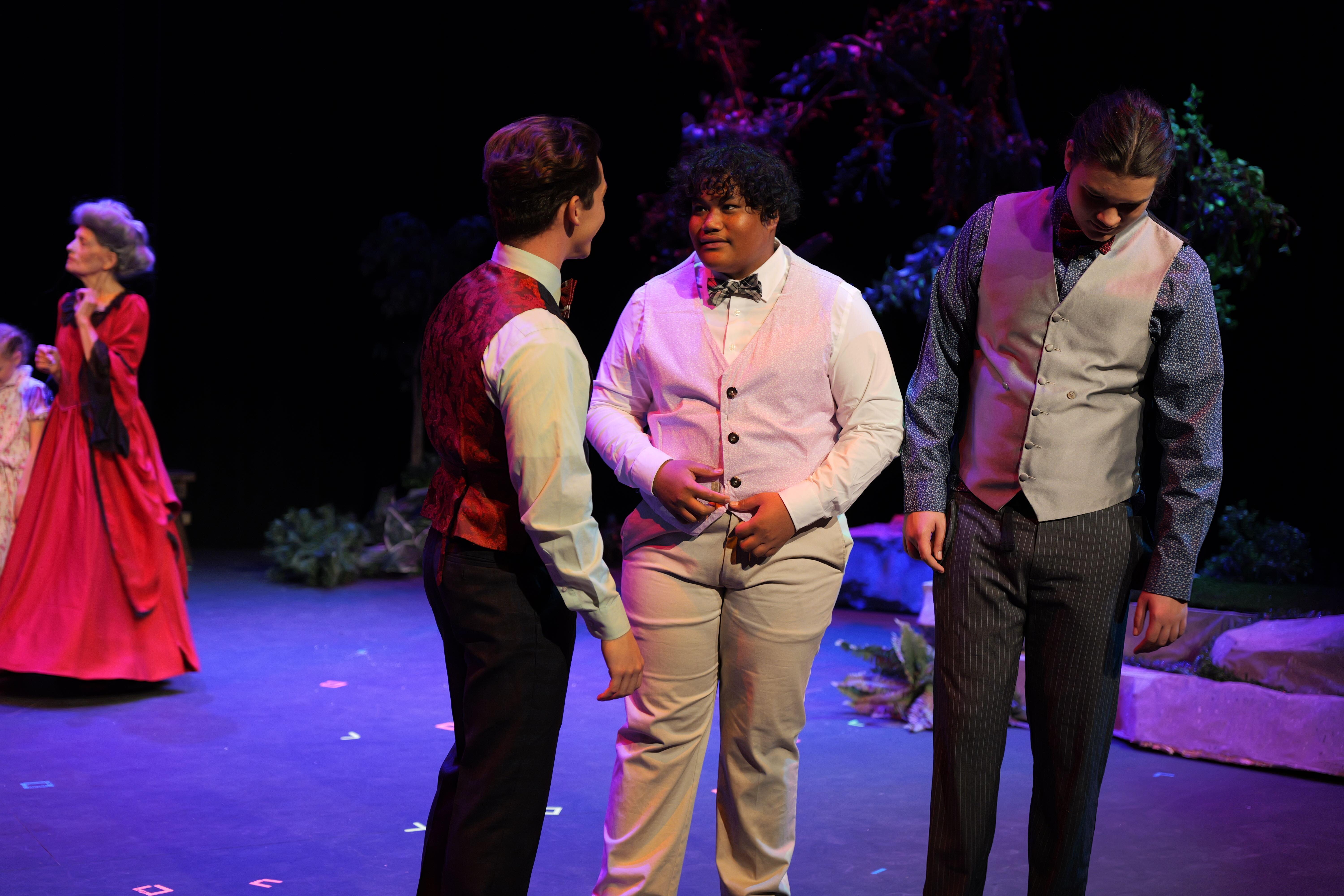 Three individuals in formal outfits interact on stage under purple lighting during a performance.
