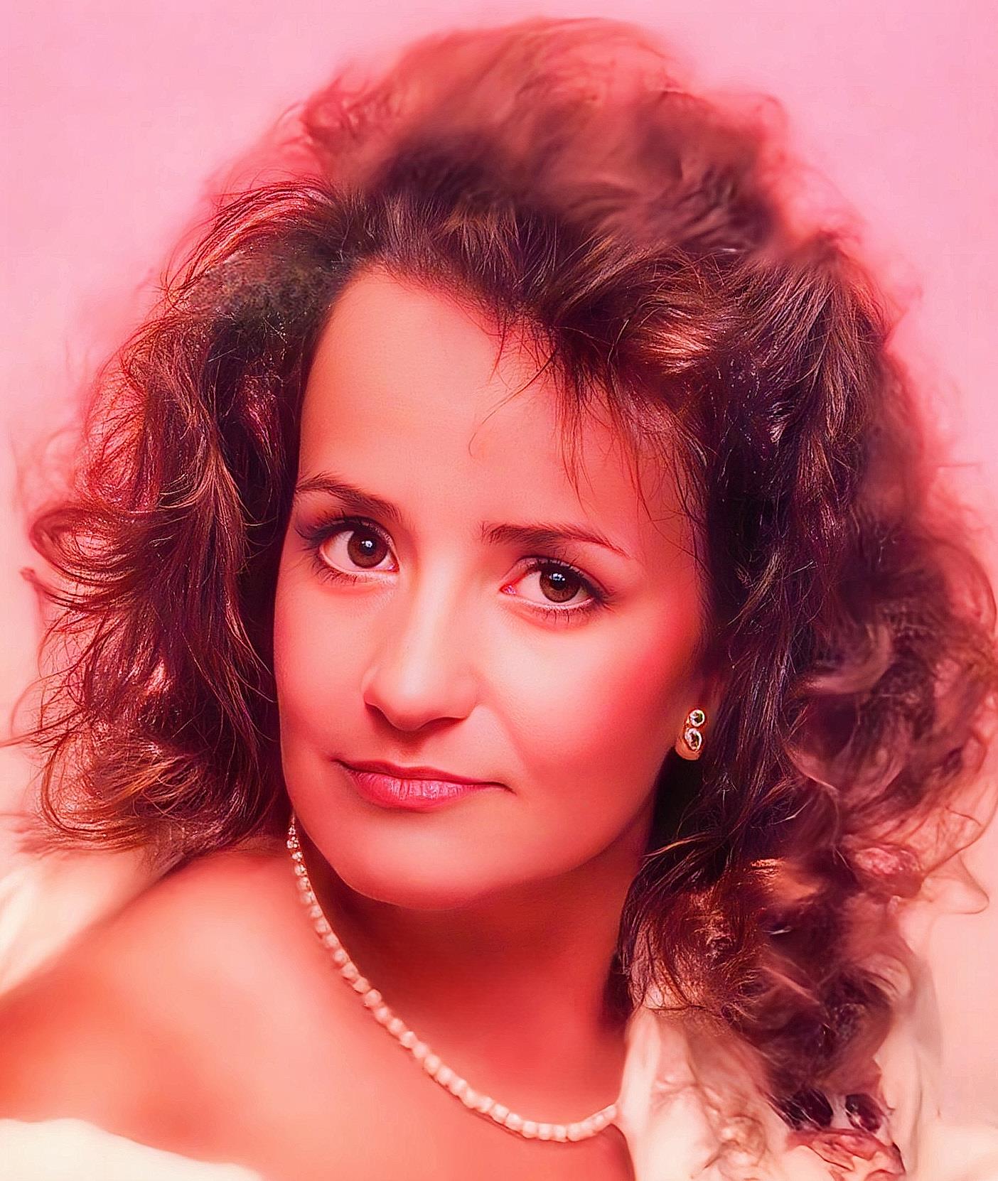 A woman stands with curly hair and a pearl necklace in a studio against a pink backdrop.