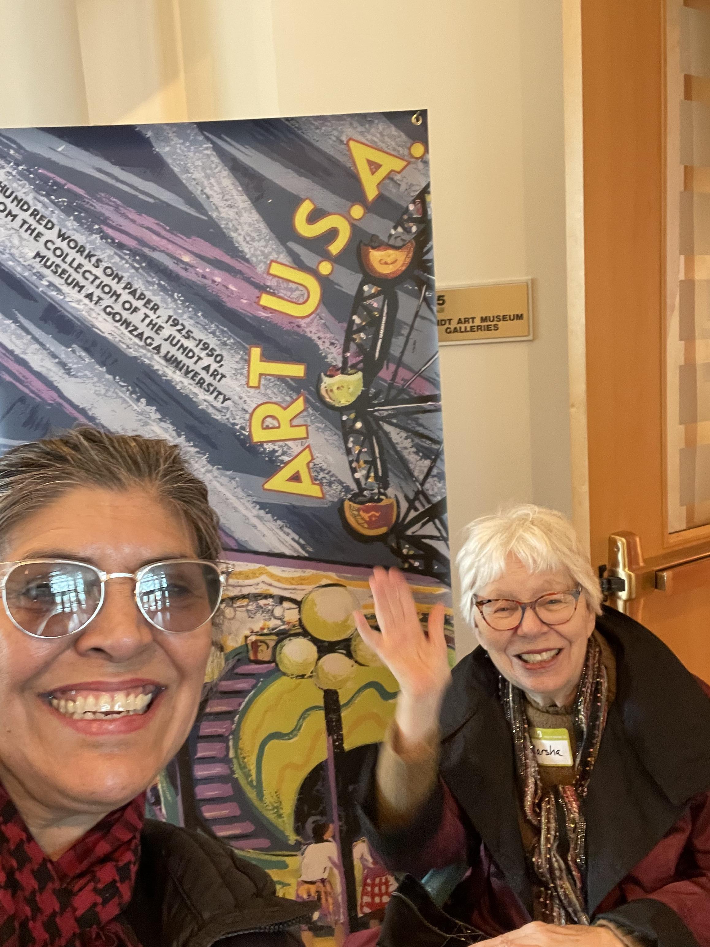Two women smile and wave in front of a creative backdrop at an art exhibit.