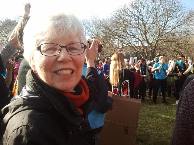 Excited older woman waves at the rally crowd, enjoying the community event in a park.