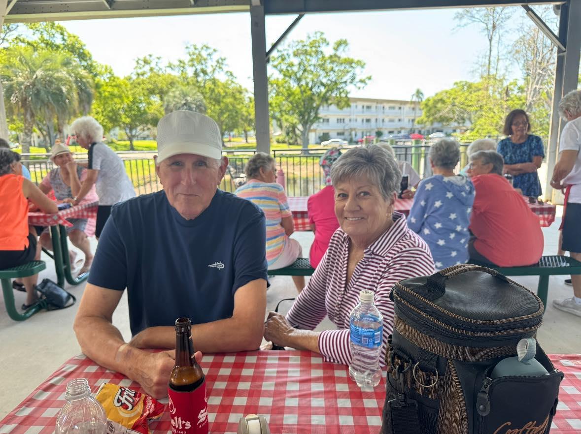 Two people sit together at a picnic table, surrounded by a lively group on a sunny day.