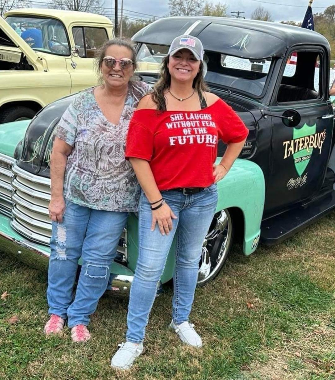 Two friends stand together beside a vintage truck, smiling and enjoying the car show atmosphere.