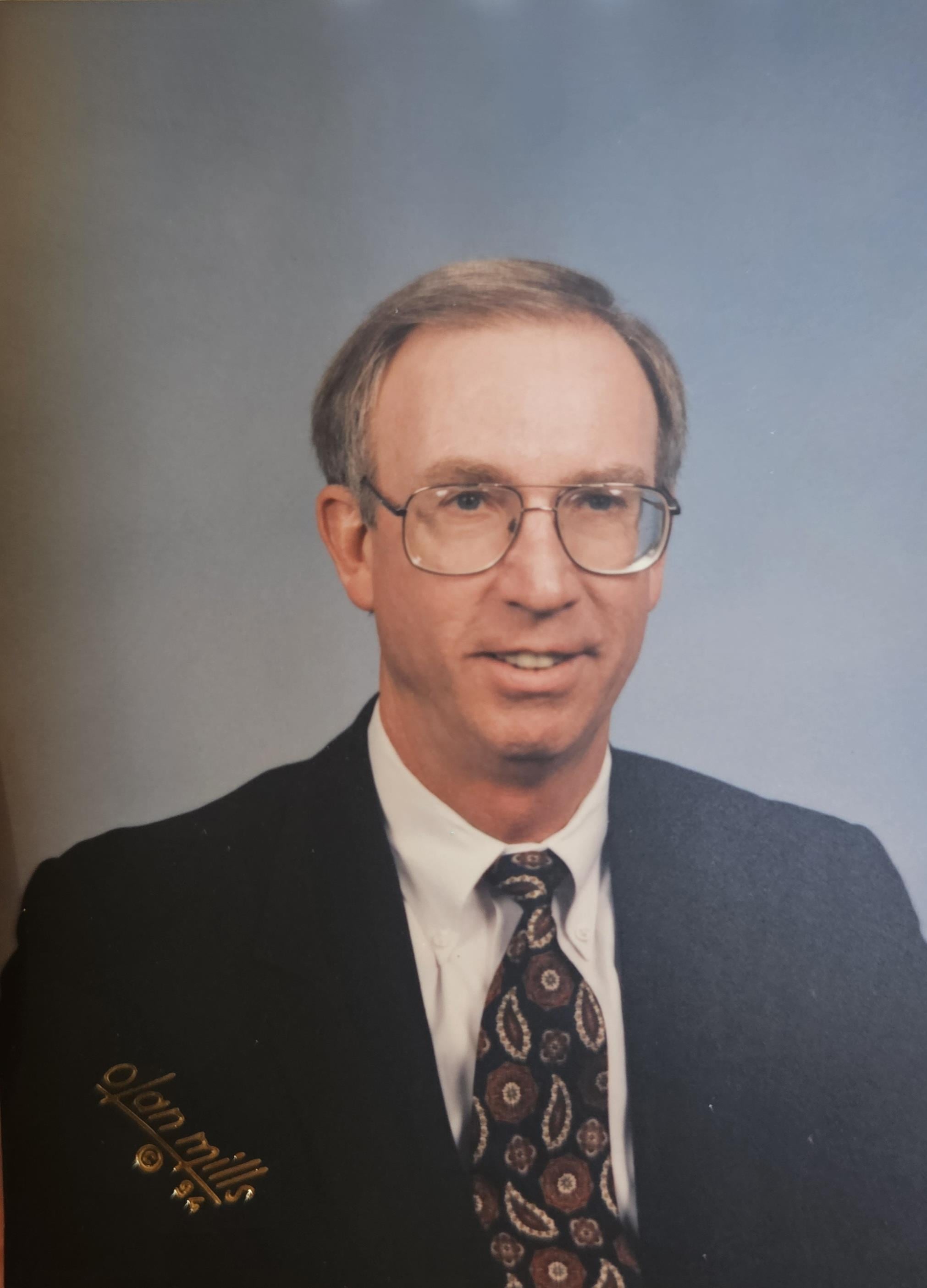 A man in formal attire stands with a slight smile, showcasing his glasses and professional demeanor.