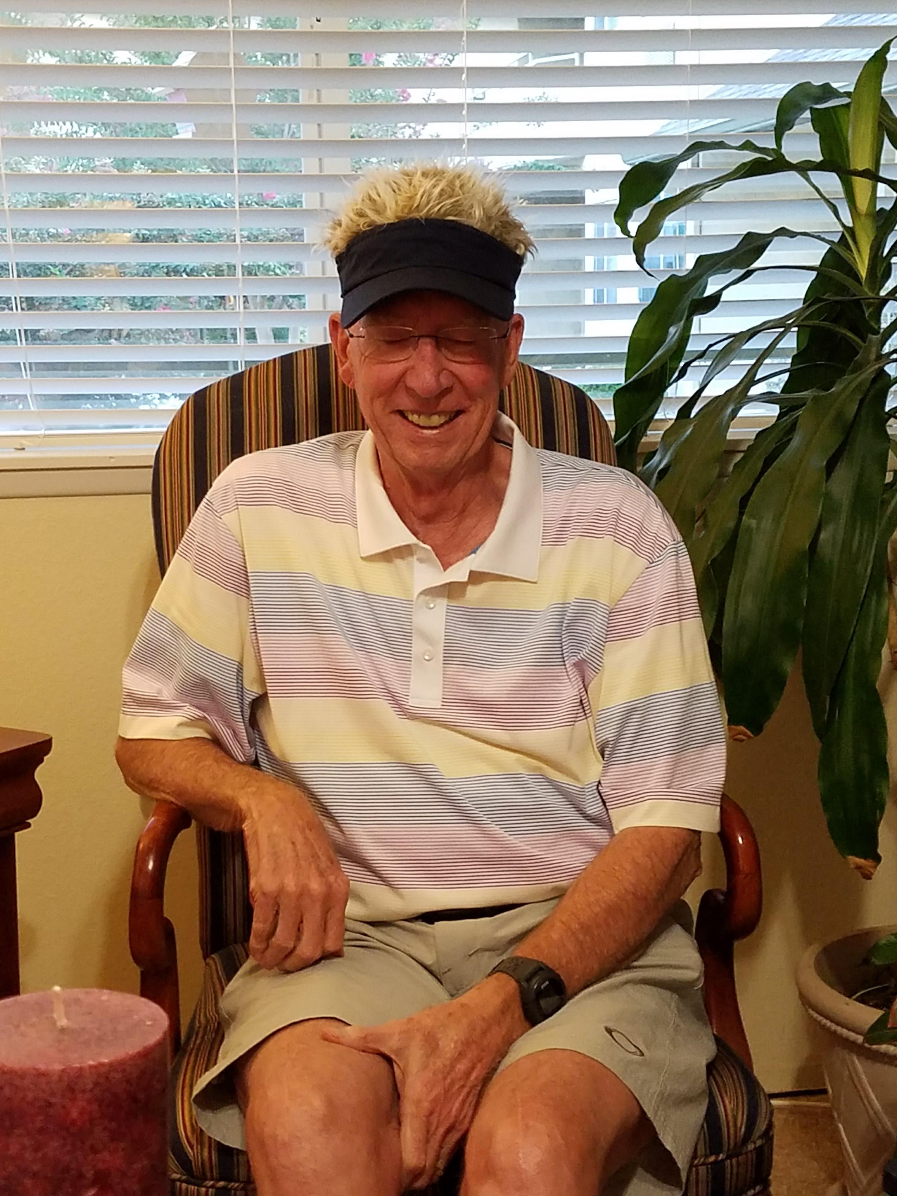 A cheerful older man relaxes in a chair wearing a visor and enjoying a moment indoors.