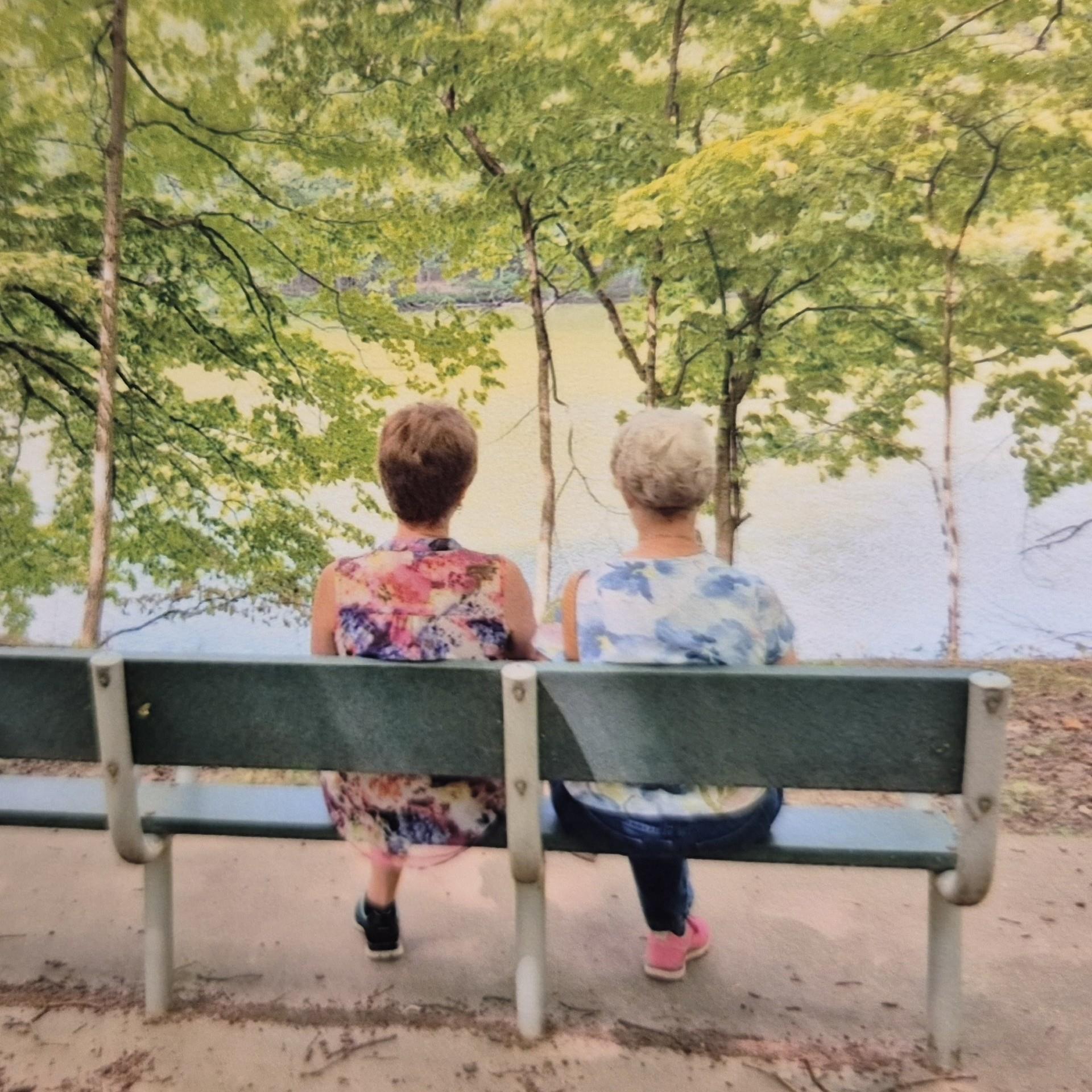 Two women relax on a bench facing a serene lake surrounded by lush greenery.