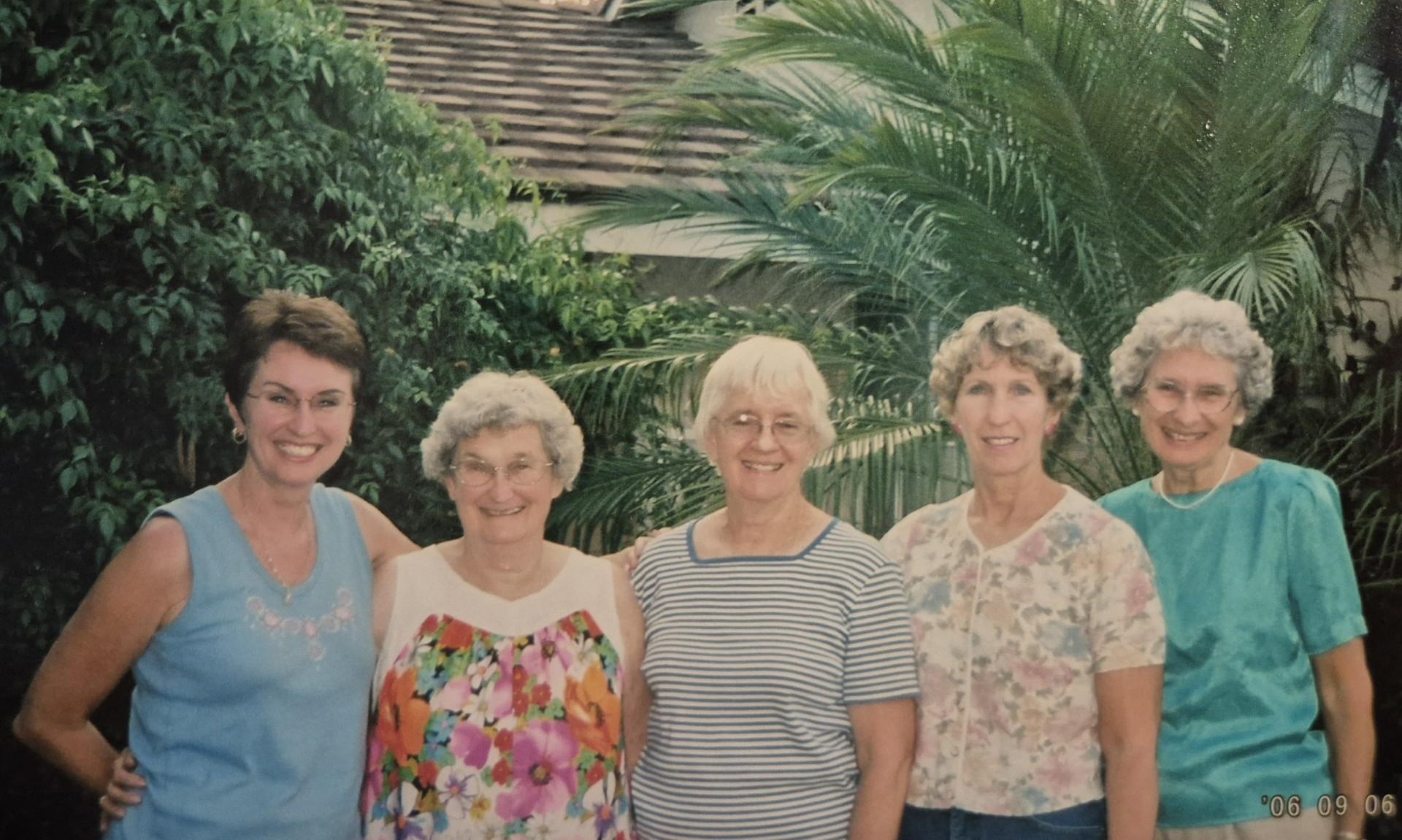 Five friends stand side by side, enjoying a sunny day in a vibrant garden with greenery surrounding.