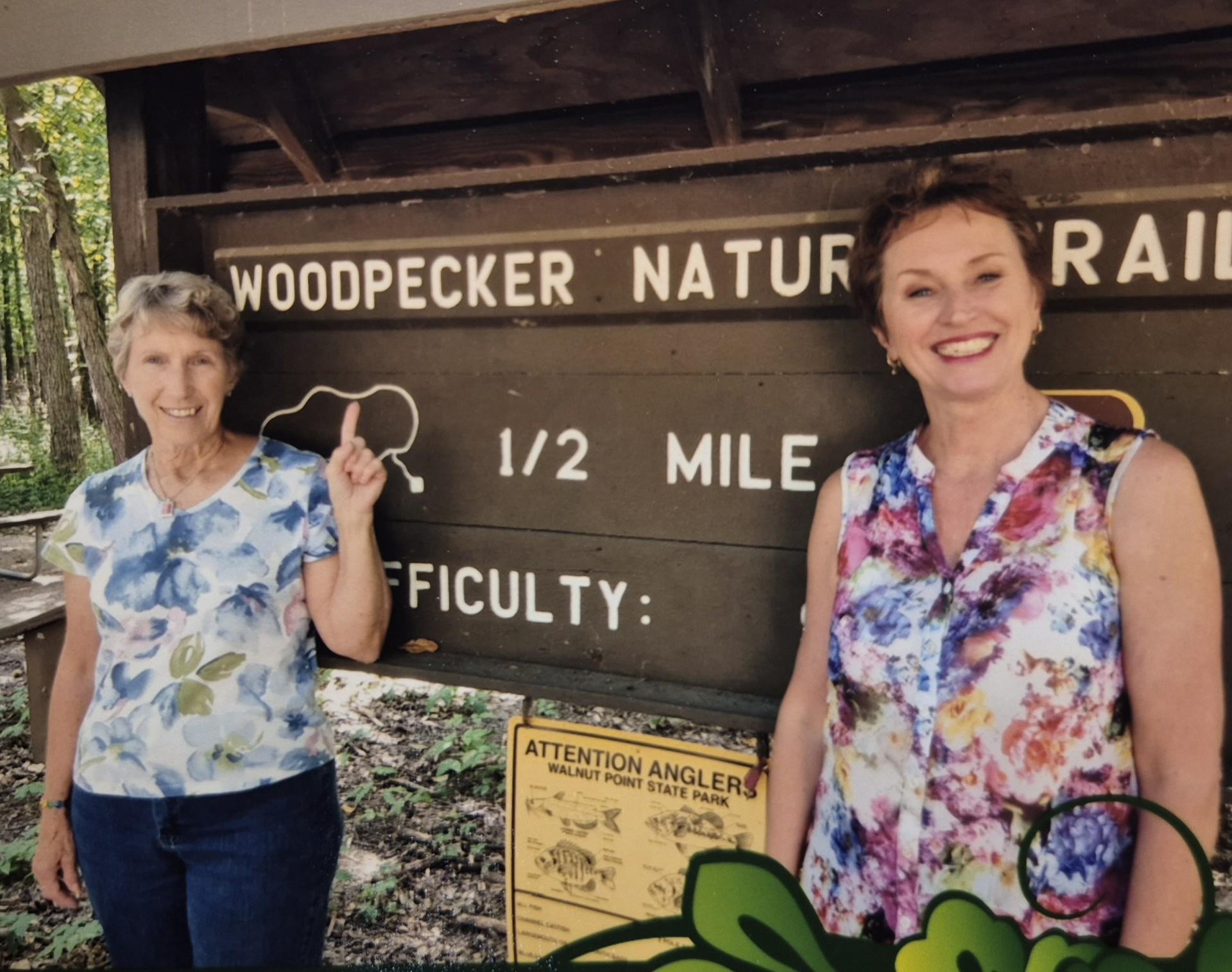 Two women are happily posing by a nature trail sign that indicates a half-mile hike.