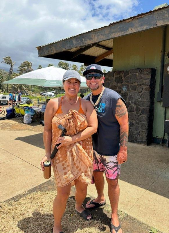 A smiling couple poses for a picture outdoors, surrounded by friends and beach activities.