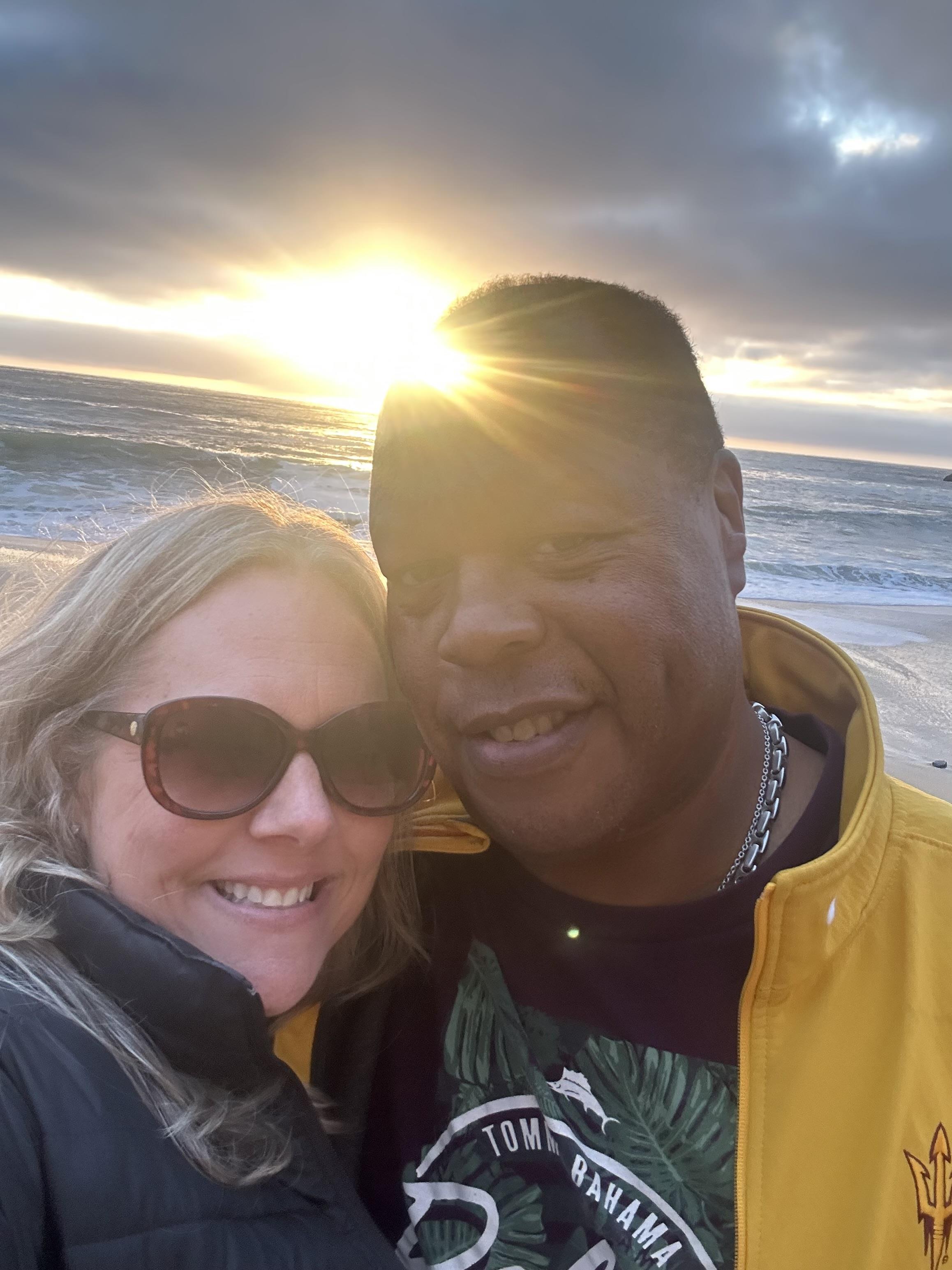 Smiling couple poses for cheerful selfie with golden sunset in the background on the beach.