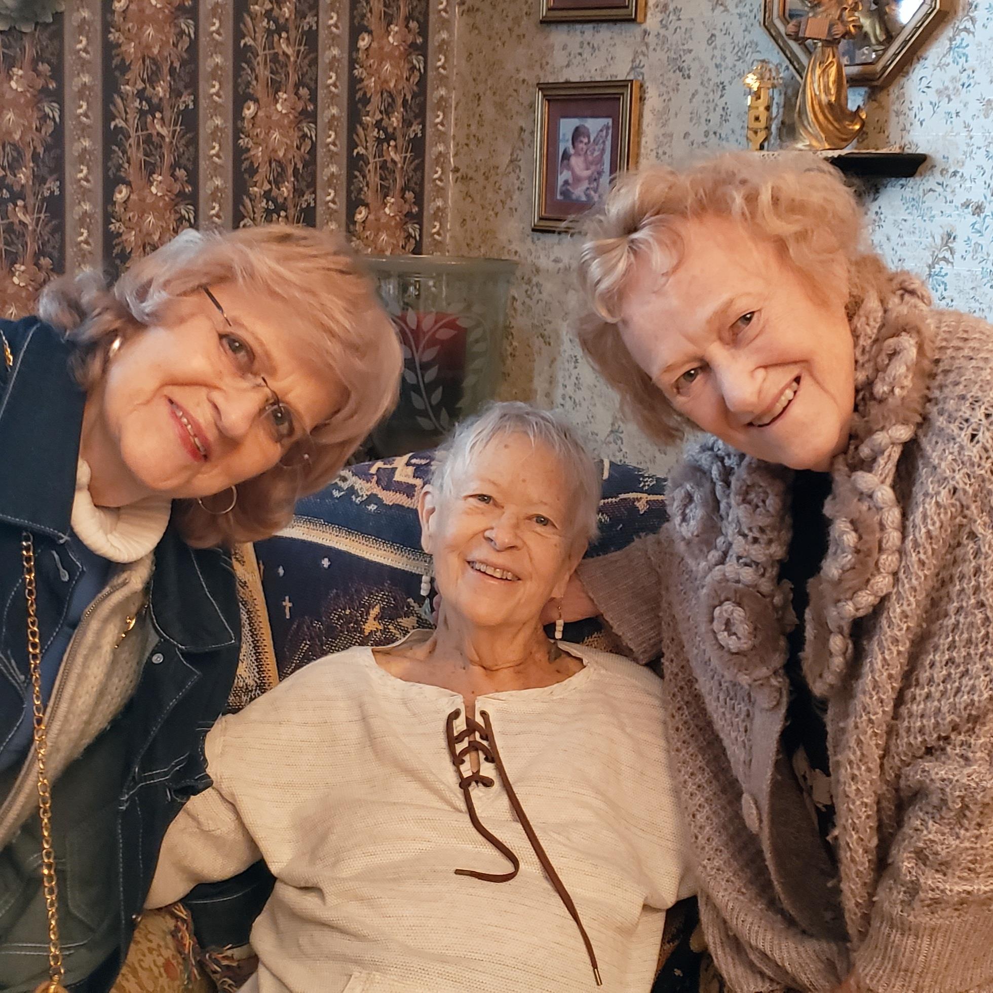 Three elderly women smile and pose together in a warm, inviting living room setting.