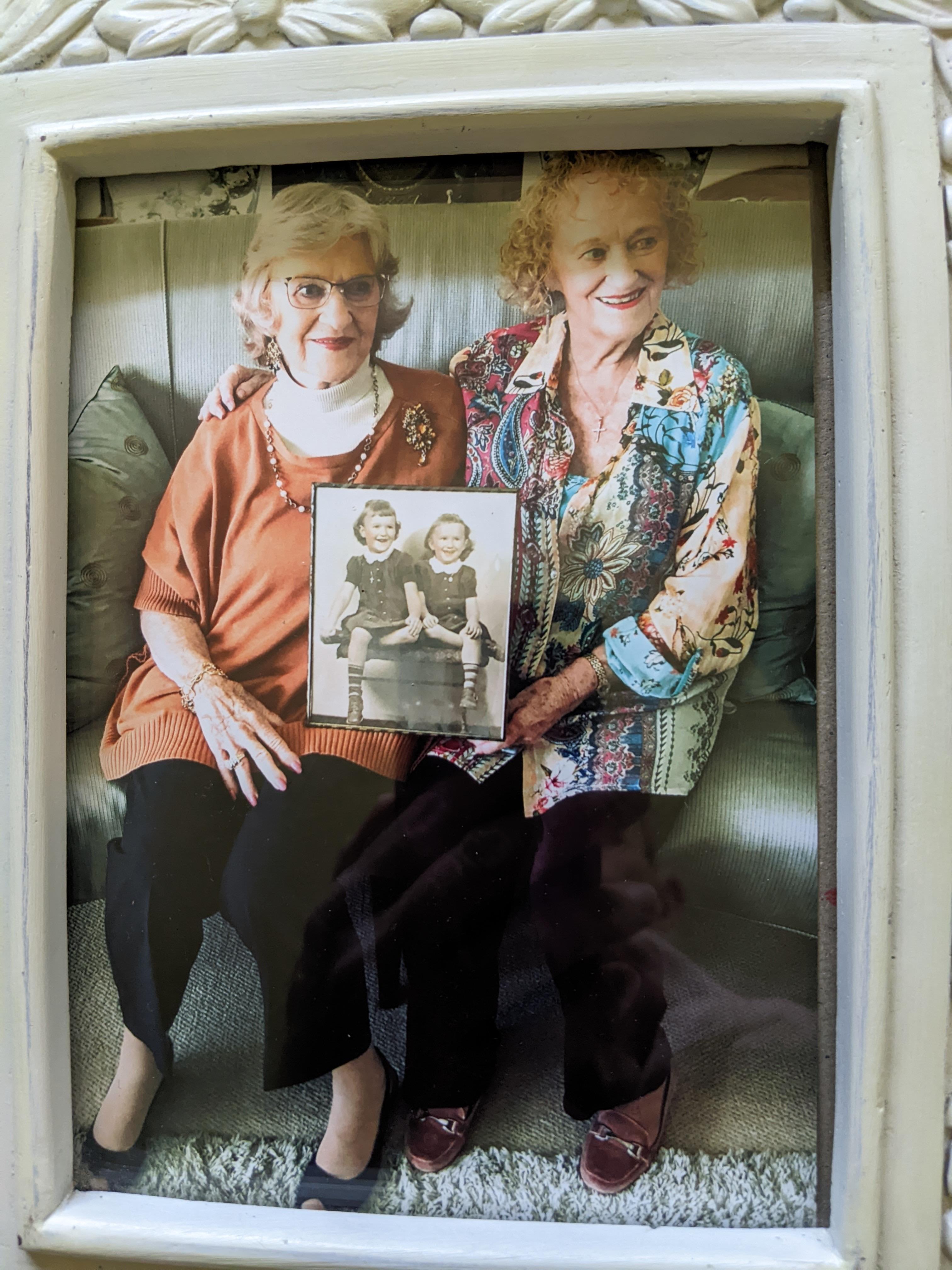 Two women sit closely on a sofa, holding a black-and-white childhood photo of themselves together.