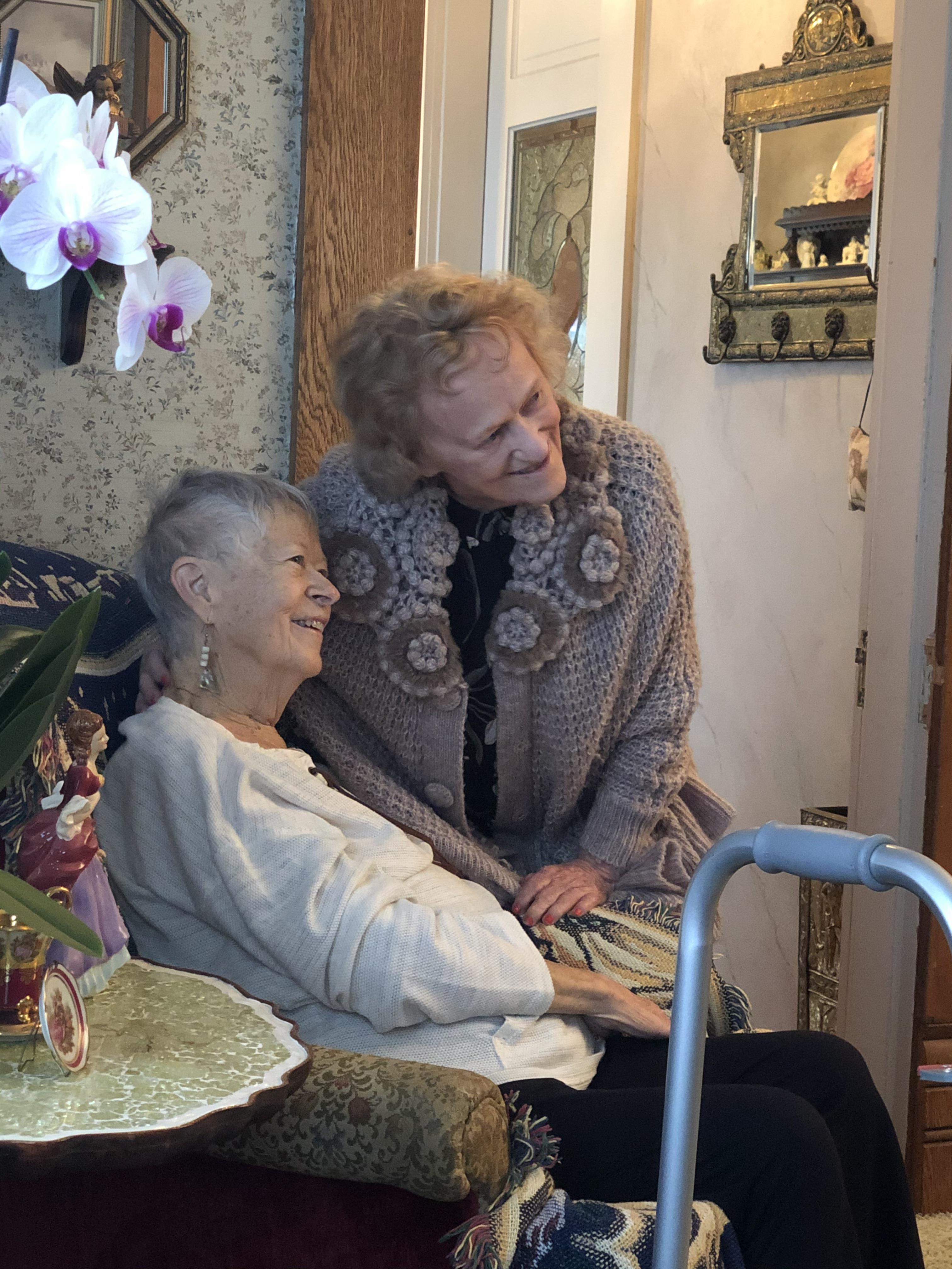 Two older women share a joyful moment in a warm and inviting living room adorned with flowers.